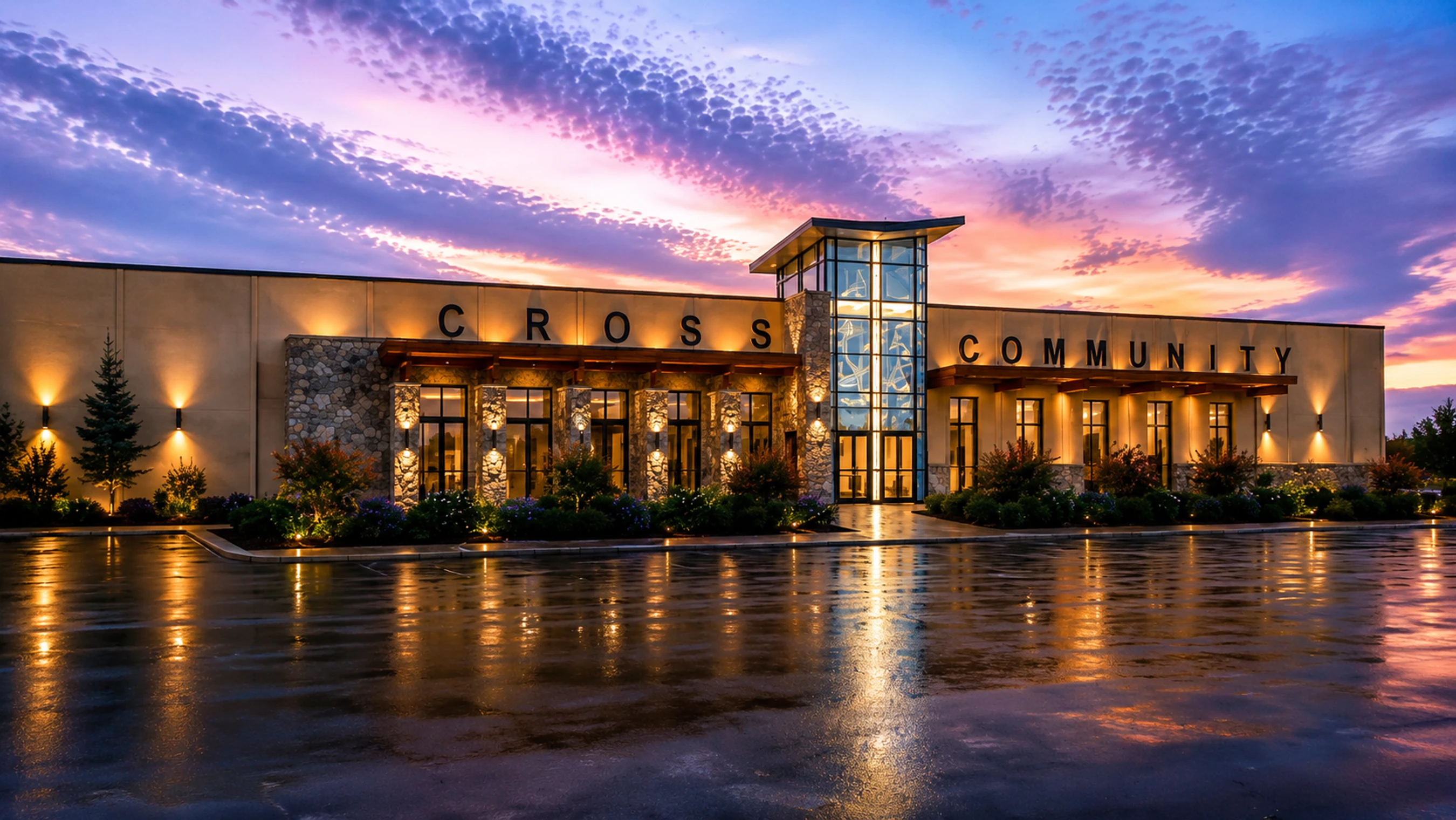 The exterior of the Cross Community building at dusk under a vibrant purple and pink sky, with its lights reflecting on the wet pavement.