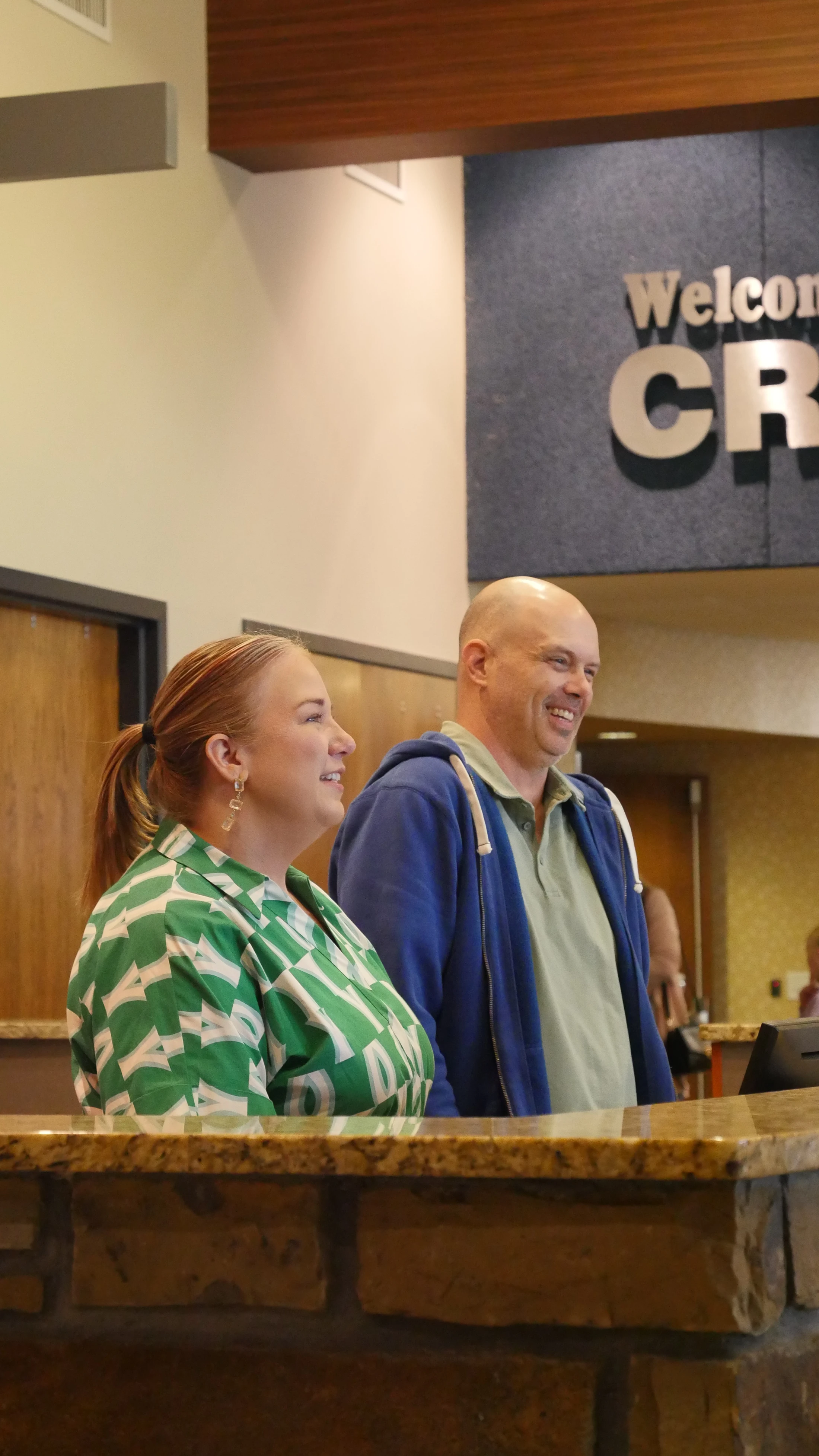 A smiling woman in a green patterned shirt and a smiling bald man in a blue hoodie stand at a stone reception desk in a lobby.