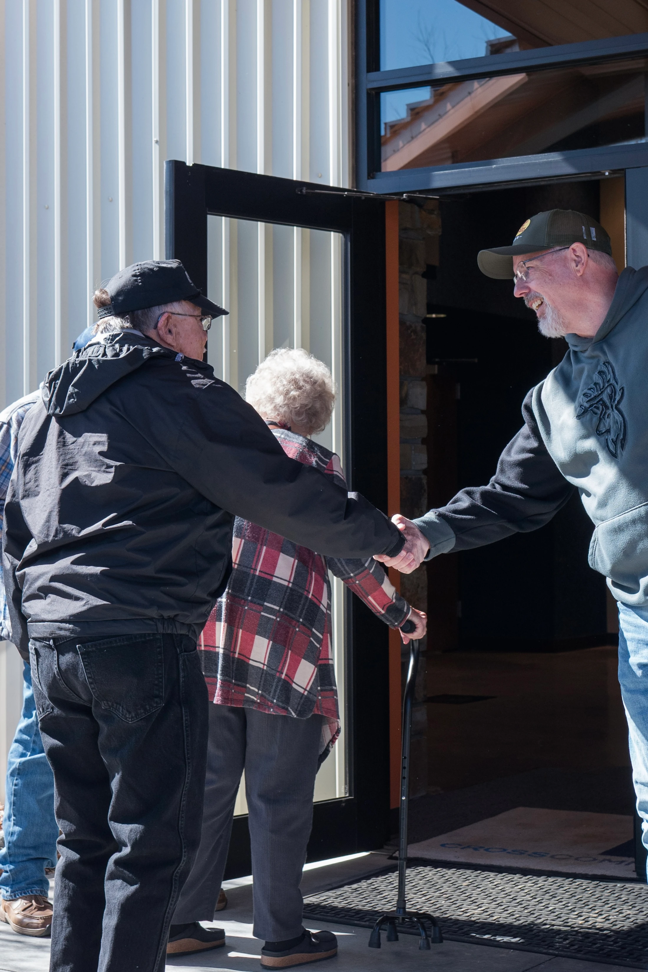 A man in a grey hoodie smiles as he shakes hands with people entering a building. An older woman with a cane walks through the doorway.