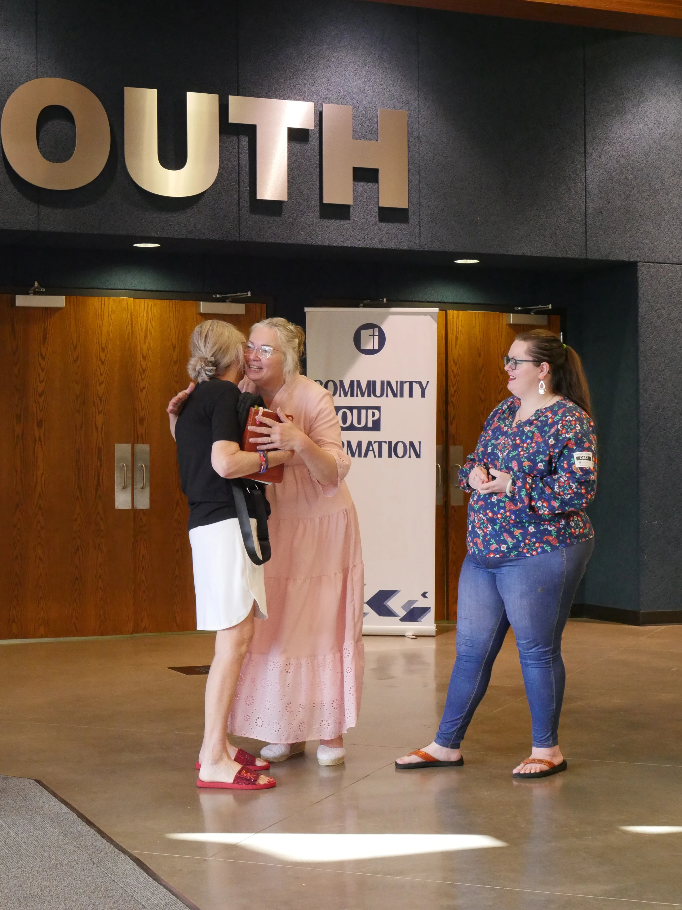 Three women in a building lobby. Two women are embracing in a warm hug while a third woman stands nearby and smiles at them.