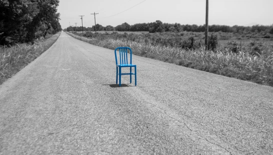A bright blue chair sits in the middle of a long, paved country road. The chair is in color while the surrounding rural landscape is in black and white.