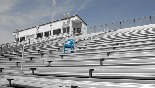 A low-angle shot of empty, metal stadium bleachers in black and white, with a single bright blue chair standing out in the middle.
