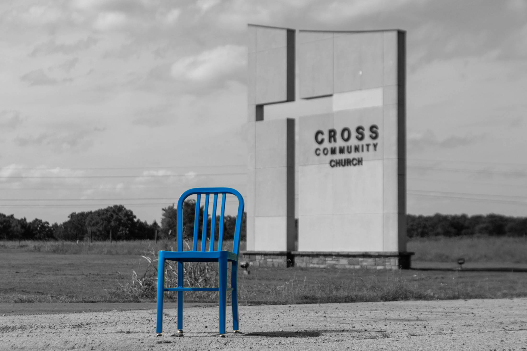 A bright blue metal chair stands on a gravel path in front of a modern church sign. The image is black and white except for the chair, which is vividly colored.