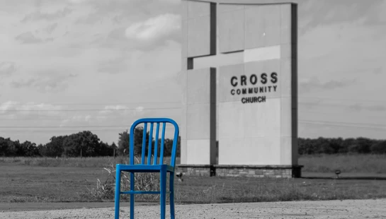 A bright blue metal chair stands on a gravel path in front of a modern church sign. The image is black and white except for the chair, which is vividly colored.