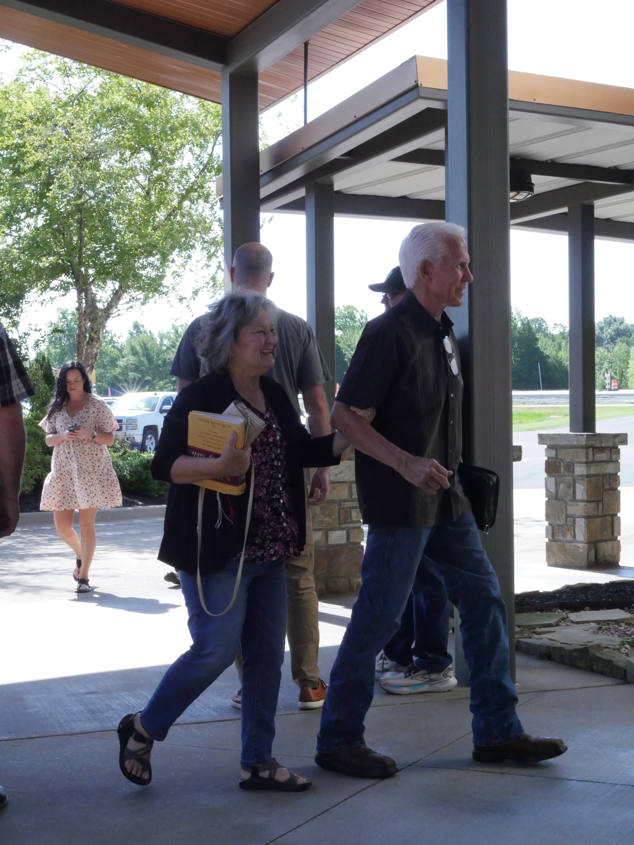 An older couple walks under a modern covered walkway. The woman smiles and holds a book, while the man walks beside her. Other people are in the background.