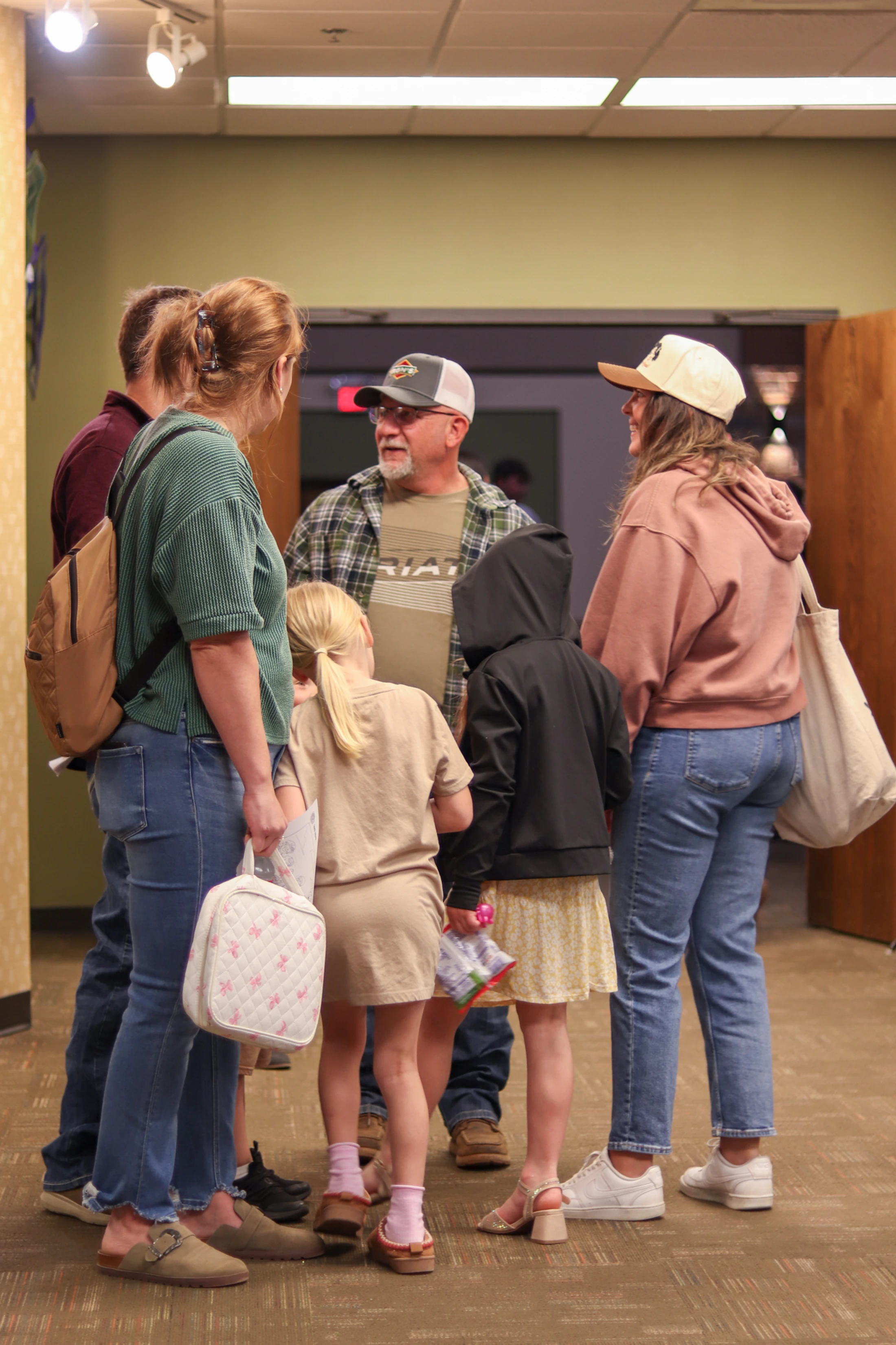 A group of several adults and two young children stand together in a circle, talking in a carpeted hallway with overhead lighting.