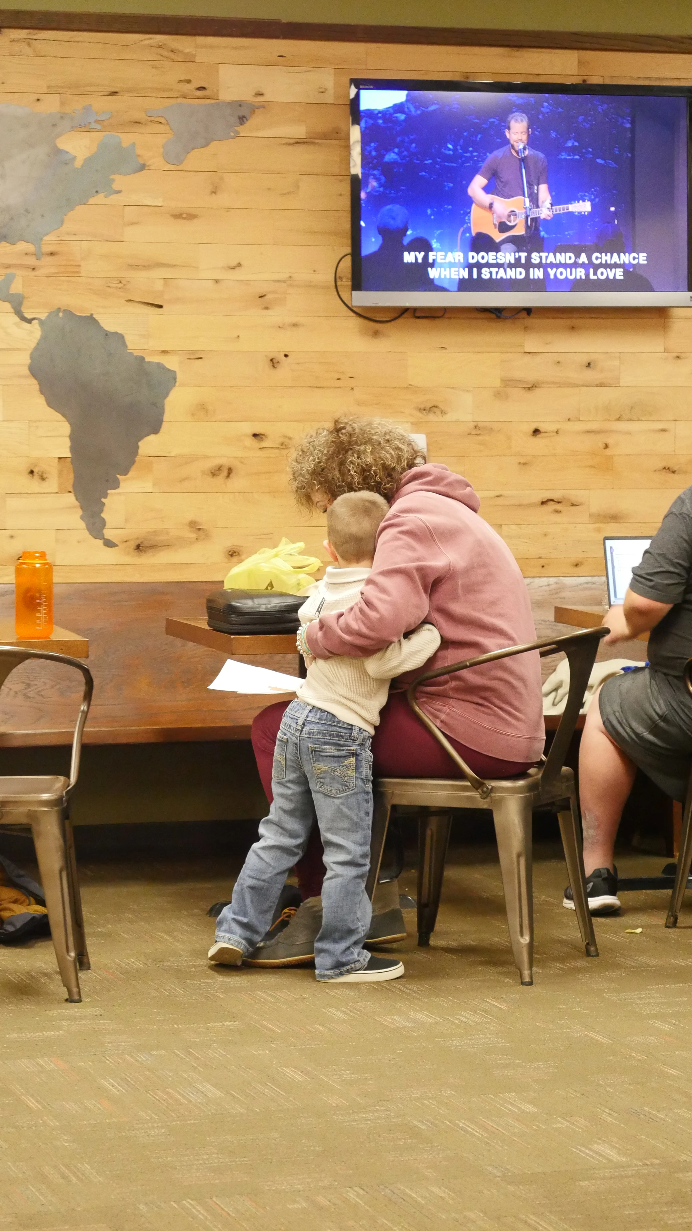 A woman with curly hair is hugged by a small boy while they watch a worship service on a TV screen. The wall behind them is wood with a world map.
