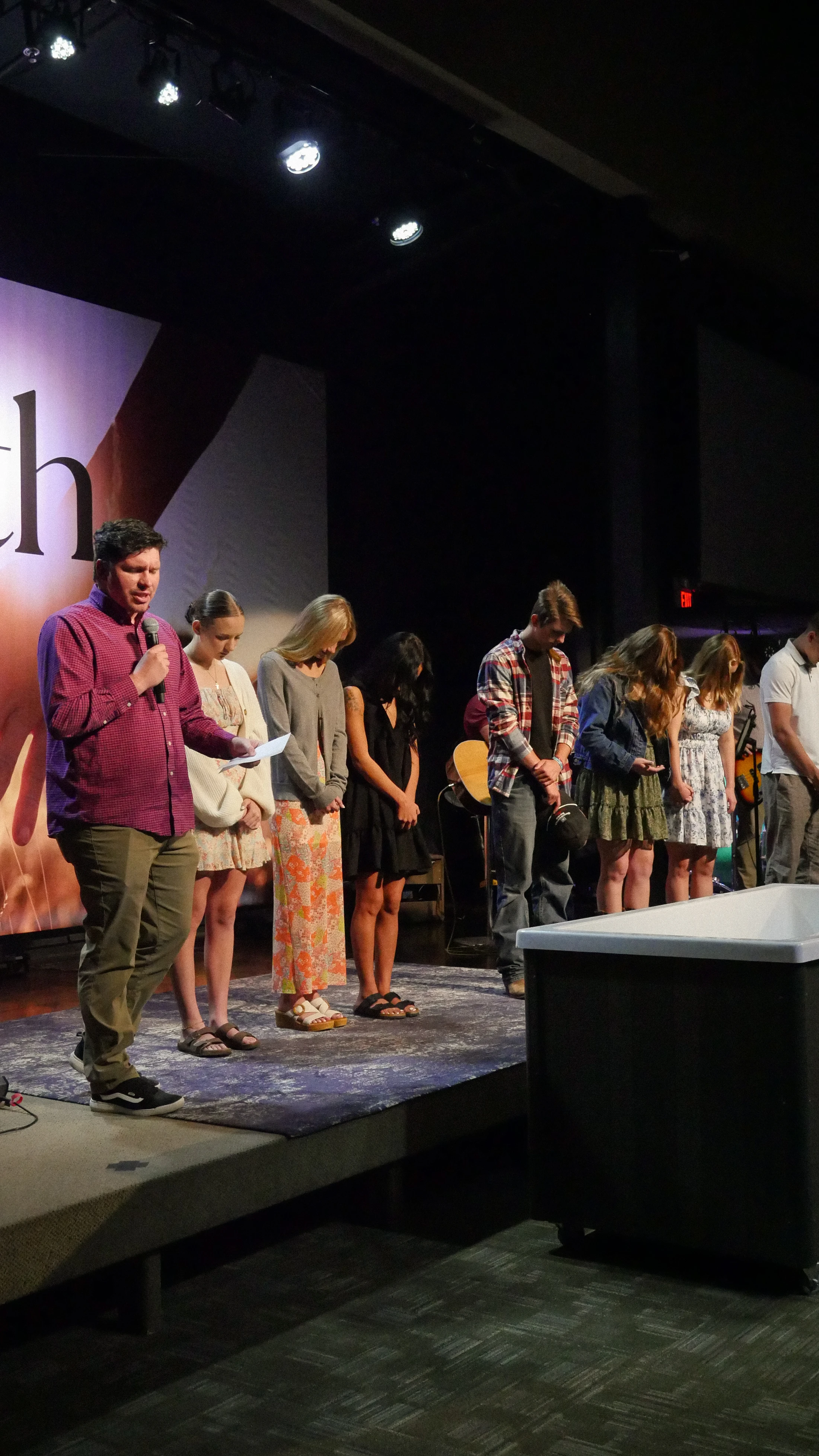 A pastor speaks into a microphone on stage while a group of people stand in a line behind him with their heads bowed, near a large white baptismal tub.