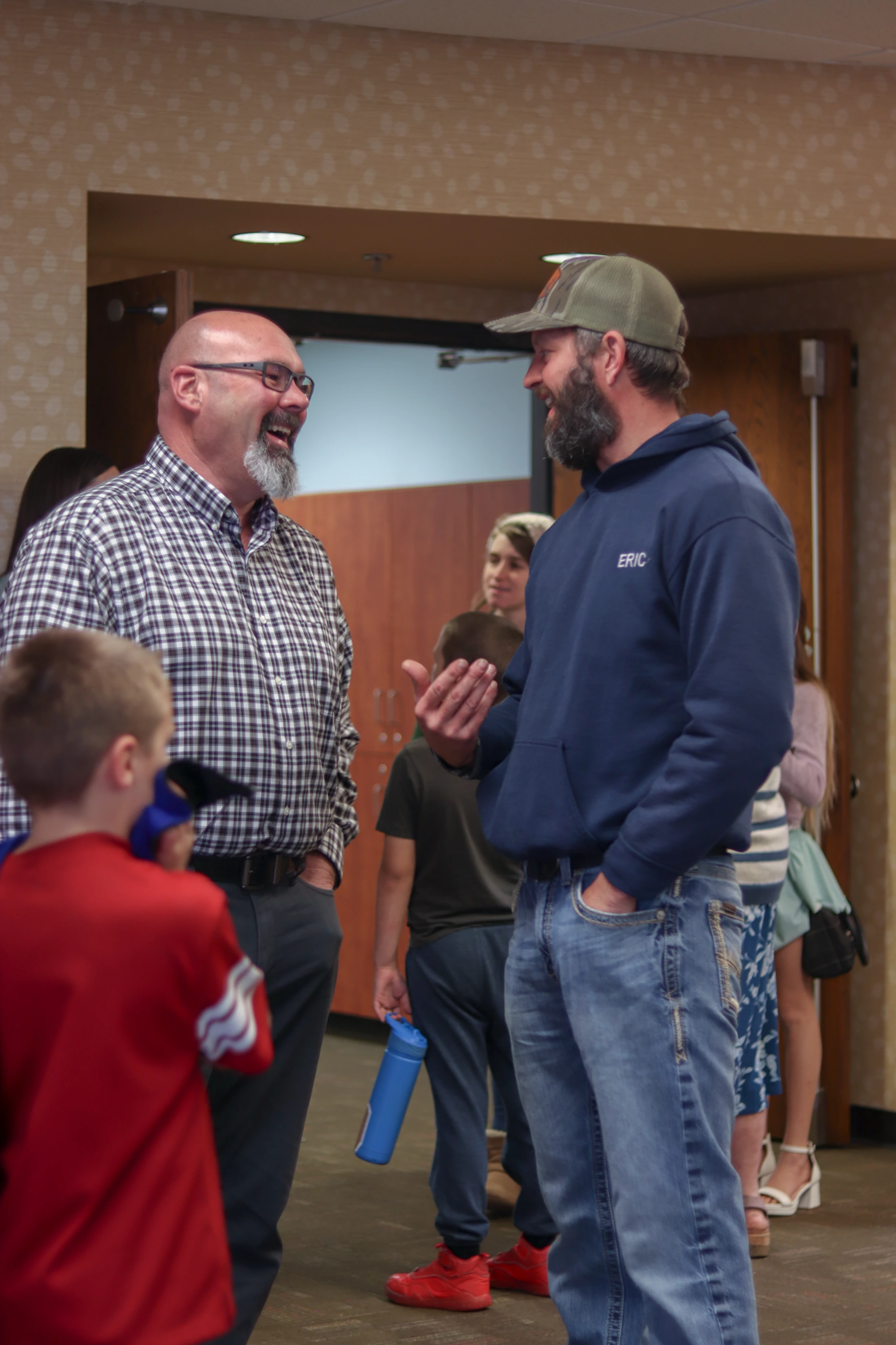 Two smiling men, one in a plaid shirt and one in a blue hoodie and cap, stand in a hallway having an animated conversation among other people.