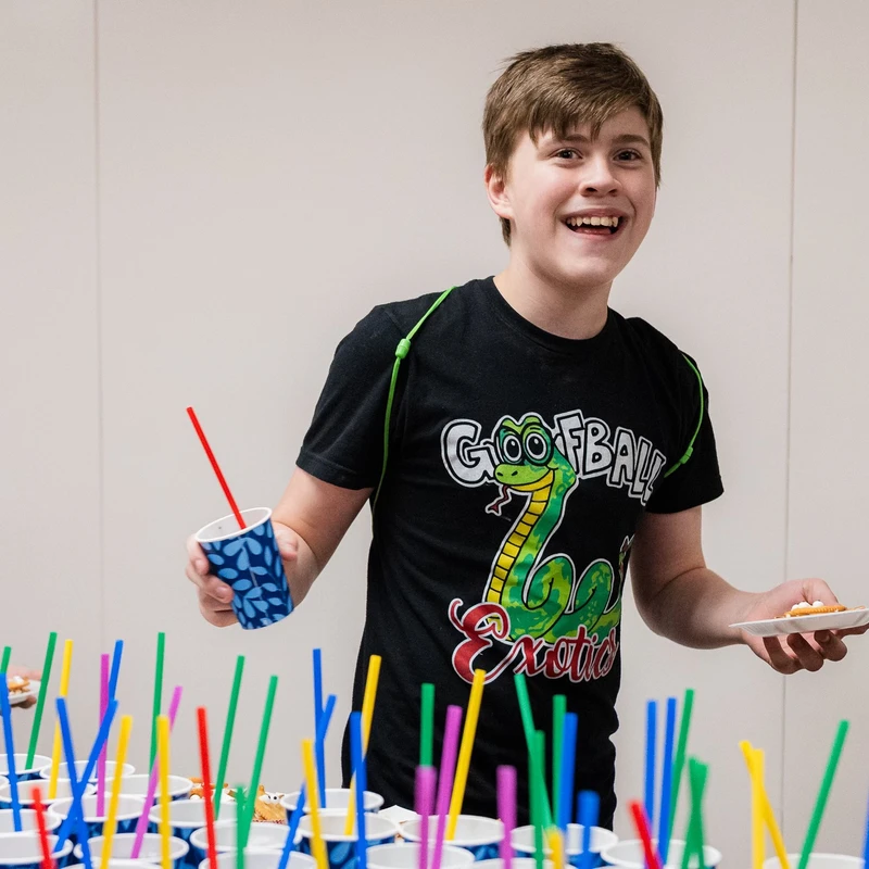 A happy boy in a black graphic t-shirt smiles while holding a cup and a plate, standing at a table full of drinks with colorful straws for a party.