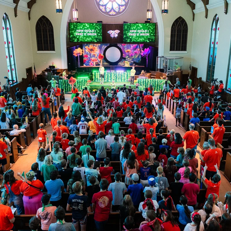 A high-angle view of a large crowd of children and adults in a church sanctuary, facing a brightly decorated stage during a worship event.