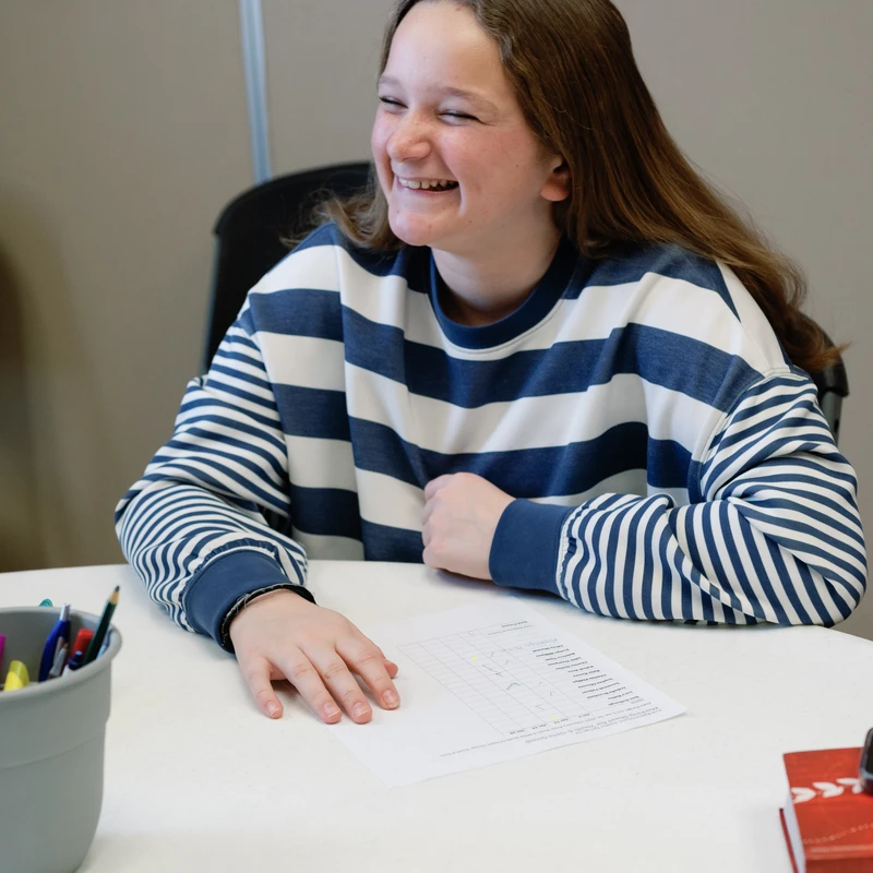 A young woman with long brown hair, wearing a blue and white striped sweater, sits at a white table and smiles while looking off to the side.