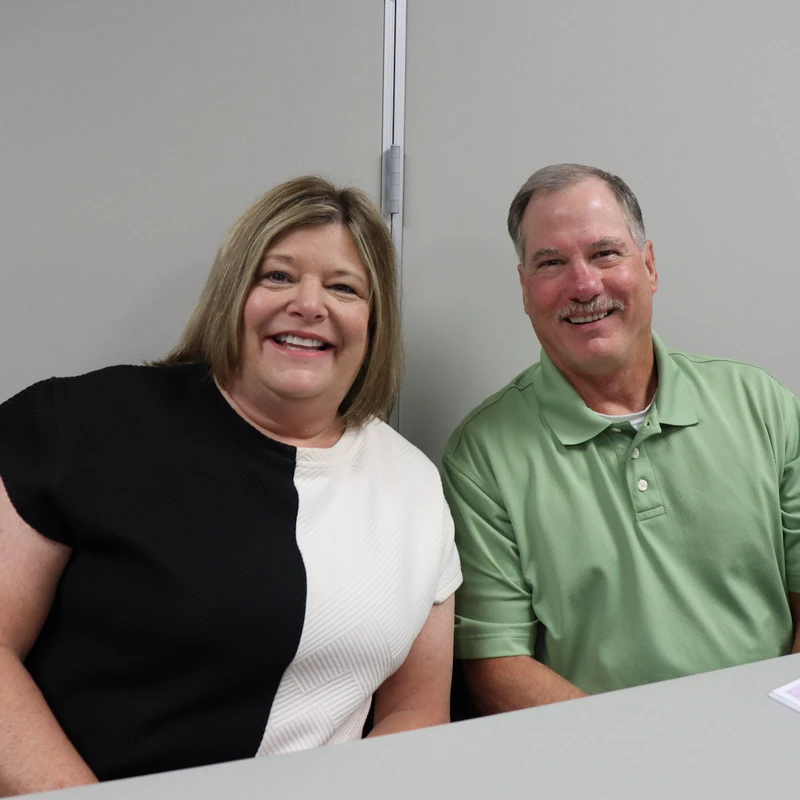 A smiling middle-aged woman in a black and white top and a man in a green polo shirt sit next to each other at a gray table, looking at the camera.