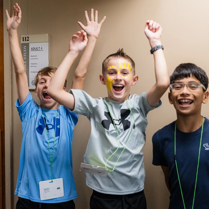 Three smiling young boys stand in a hallway, cheering with their arms in the air. The boy in the center has yellow face paint and a big, happy expression.