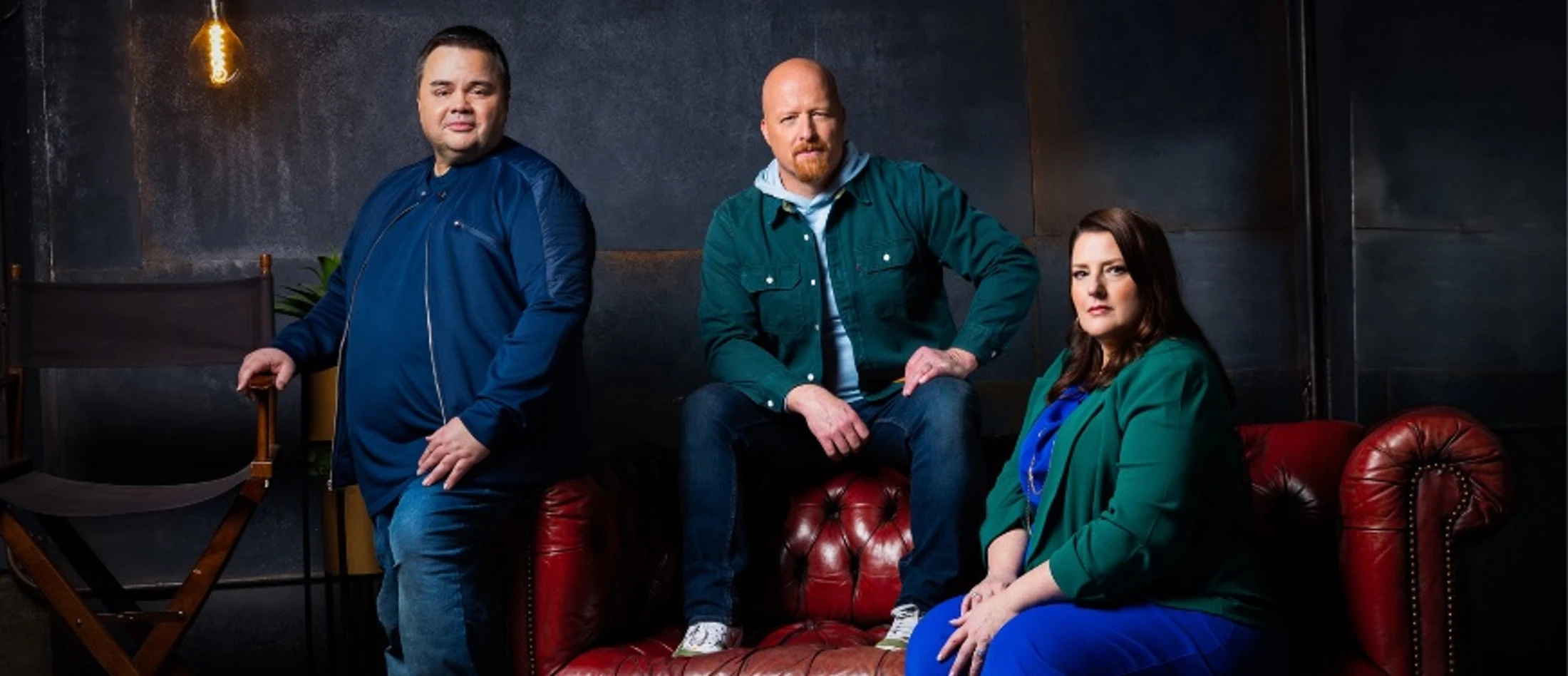 Two men and one woman posing for a professional portrait. One man stands by a chair, while a man and a woman sit on a red leather Chesterfield sofa.