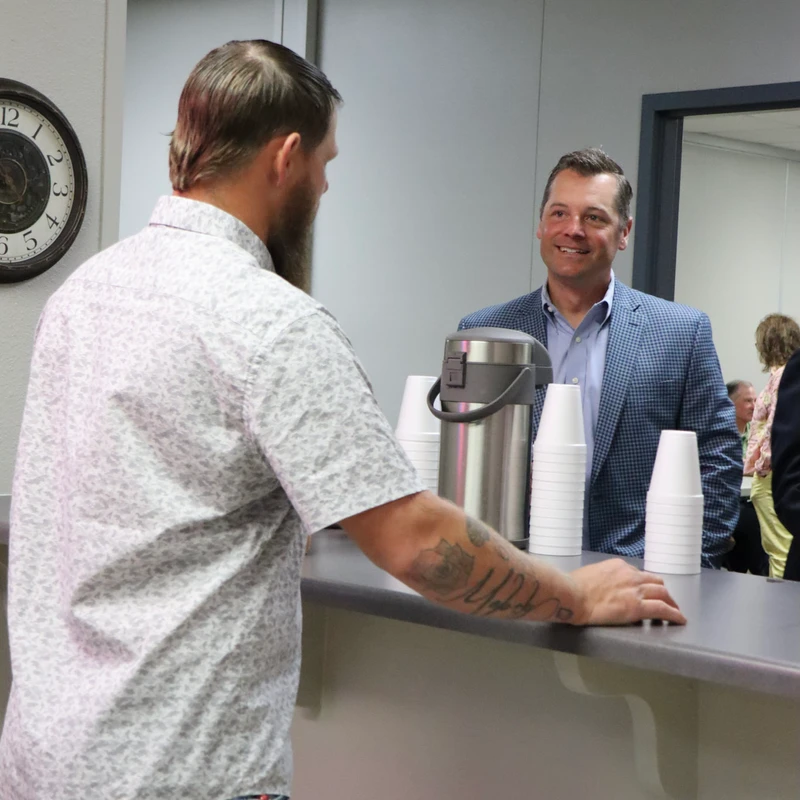 Two men chat in an office breakroom. One man with a beard stands at a counter while another man in a blazer smiles from across from him.