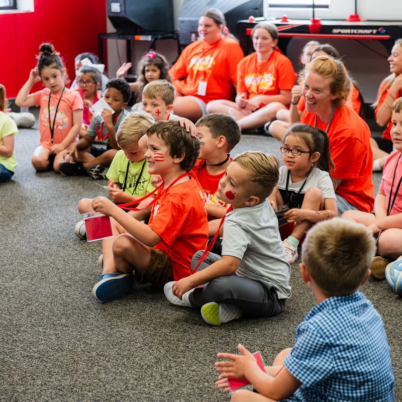 A group of elementary-aged children and several adults sit on a gray carpet, engaged in a group activity in a classroom with a red wall.