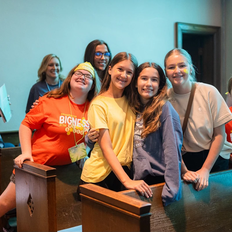 A group of smiling teenage girls and women pose for a photo together in wooden church pews. Some wear matching orange event shirts and have face paint.