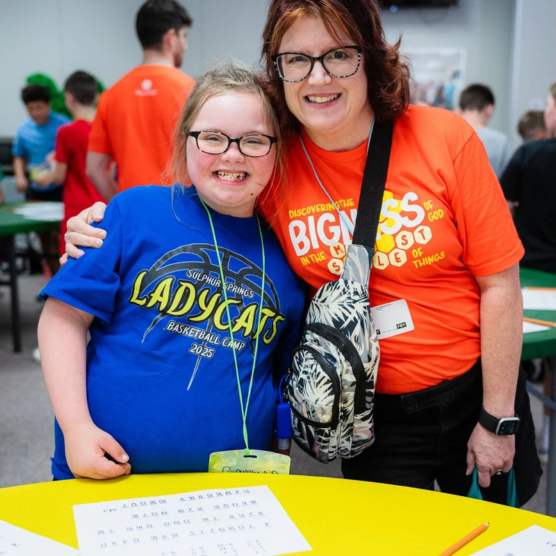 A woman in an orange shirt hugs a smiling young girl in a blue basketball camp shirt. They are both wearing glasses and posing at a yellow table.
