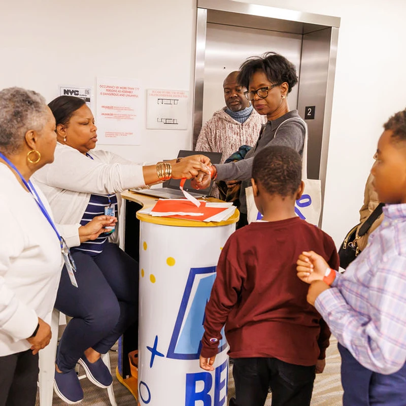 A group of Black adults and children stand at a registration desk, where a staff member puts a red wristband on an attendee's wrist.