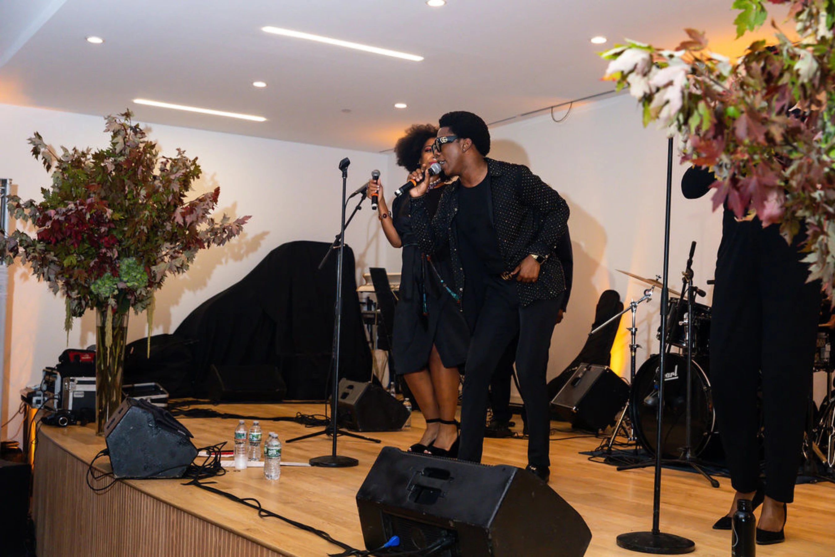 A male and a female singer in black outfits sing into microphones on a wooden stage, decorated with large autumnal floral arrangements for an event.