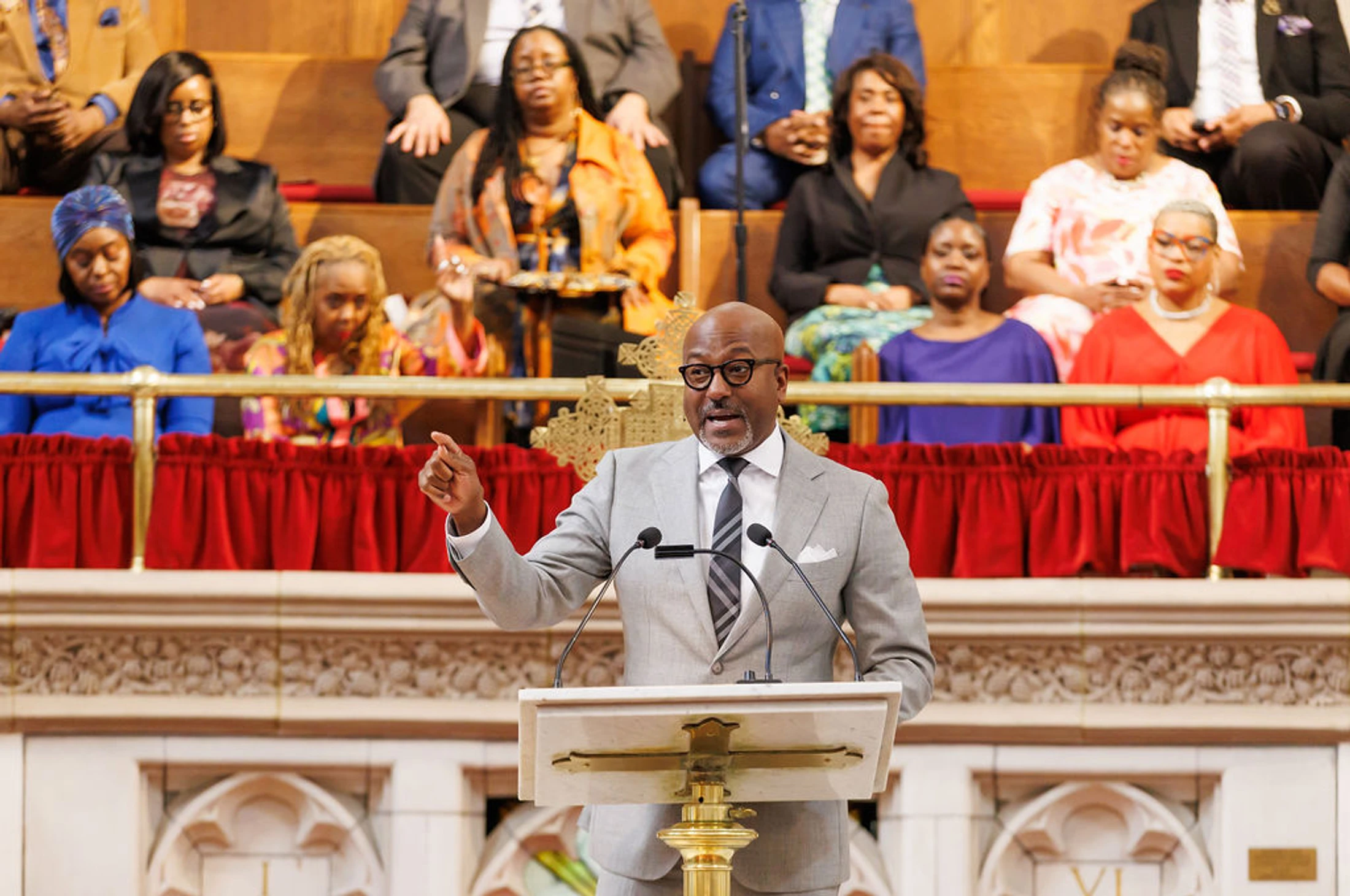A Black man wearing a grey suit and glasses speaks passionately from a podium in a church, gesturing with his finger as the congregation watches from the pews.