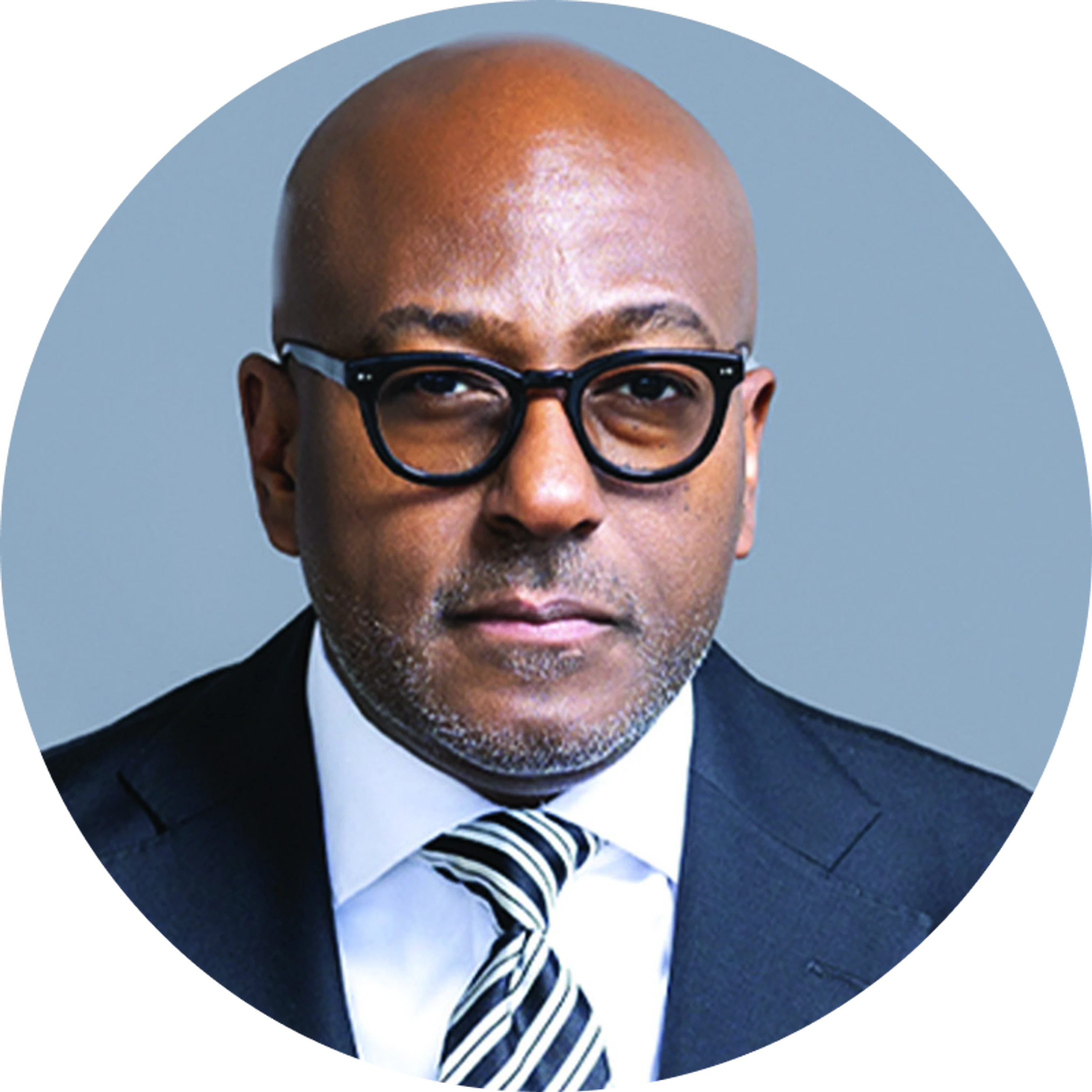 A circular headshot of a bald Black man wearing glasses, a dark suit, a white shirt, and a black and white striped tie against a gray background.
