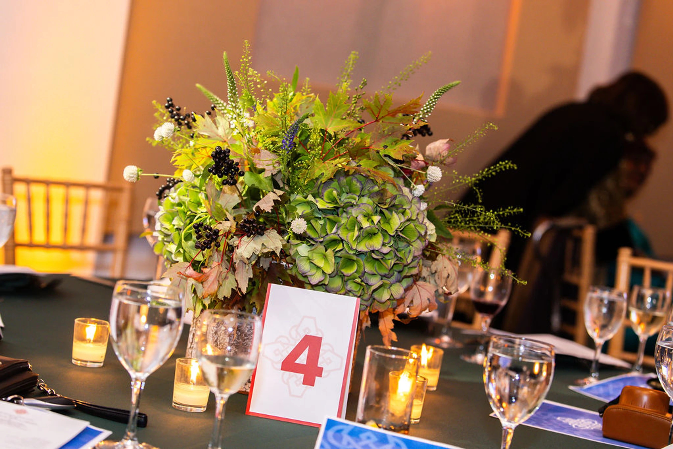 A dining table set for an event with a floral centerpiece of green hydrangeas and dark berries, next to a table number card showing a red number 4.