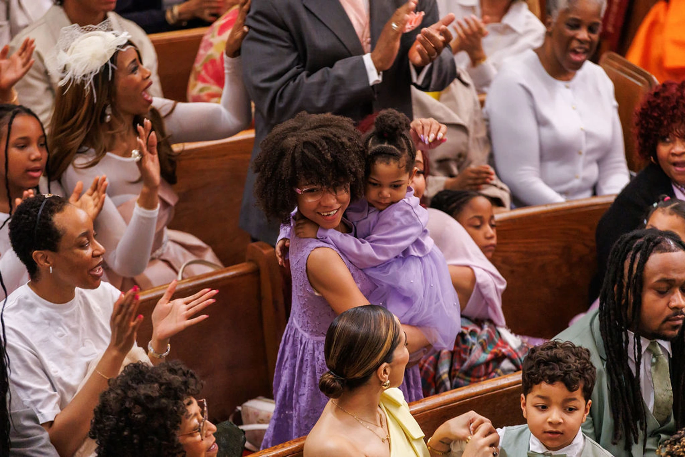 A diverse Black congregation of all ages worships in church pews. A woman in a purple dress smiles while holding a small child as others clap and sing.
