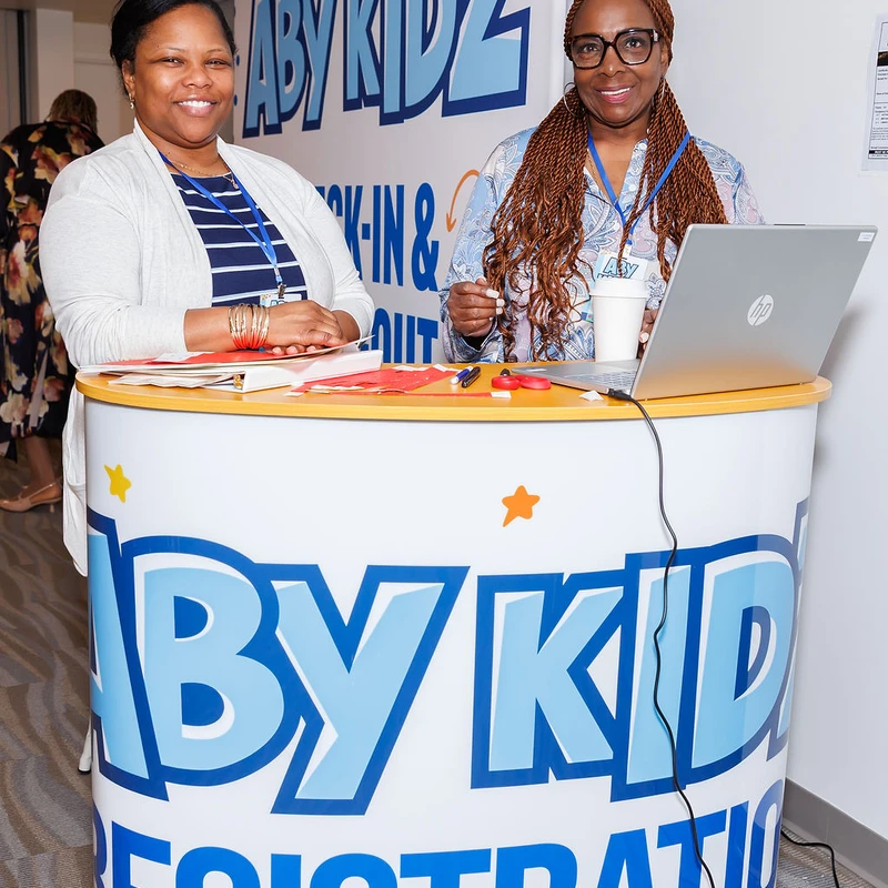 Two smiling women stand behind a colorful 'Baby Kidz Registration' desk at an event. One woman has a laptop in front of her.
