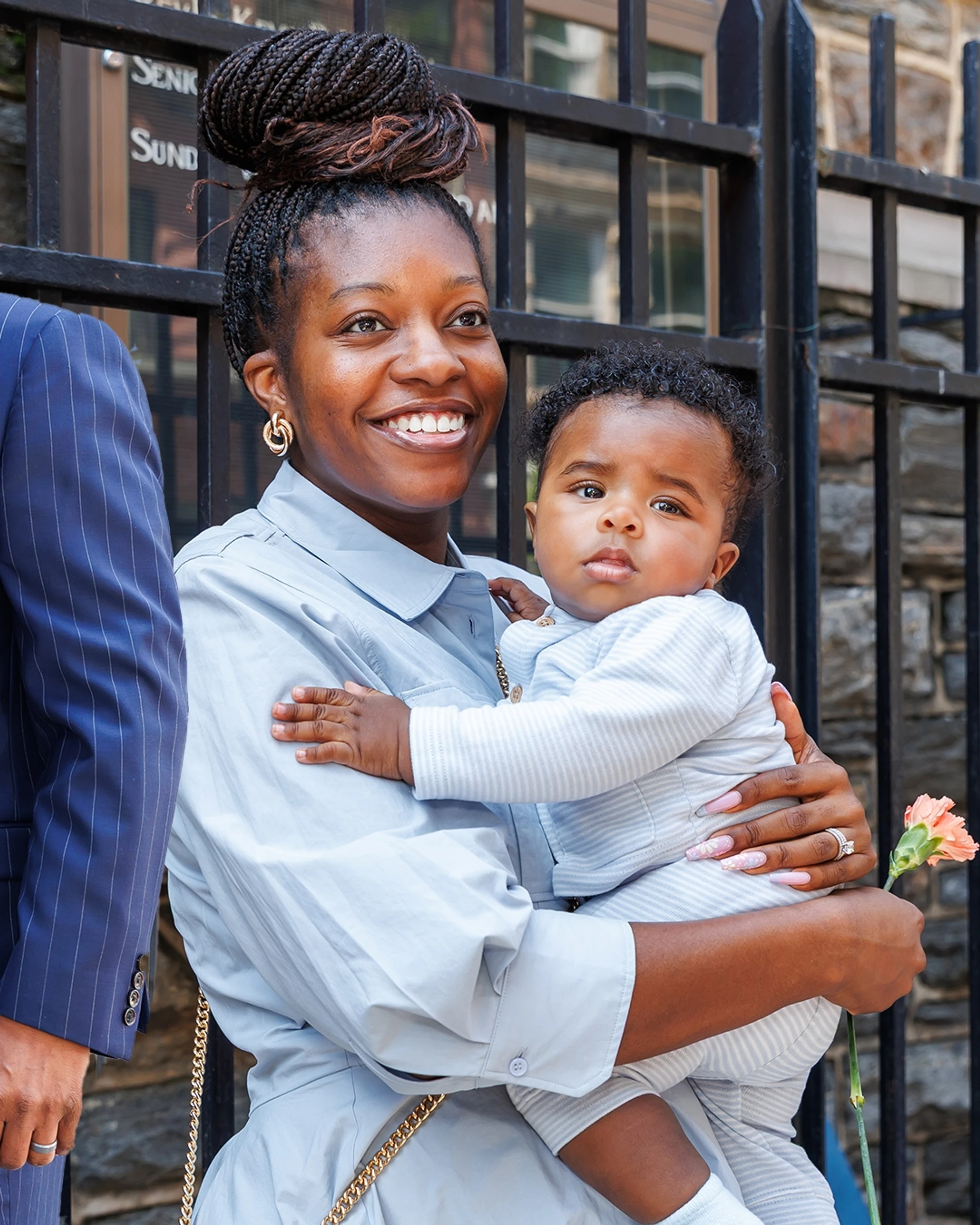 A smiling Black woman with her hair in a braided bun holds a baby in a striped outfit. They are standing outside by a black fence.