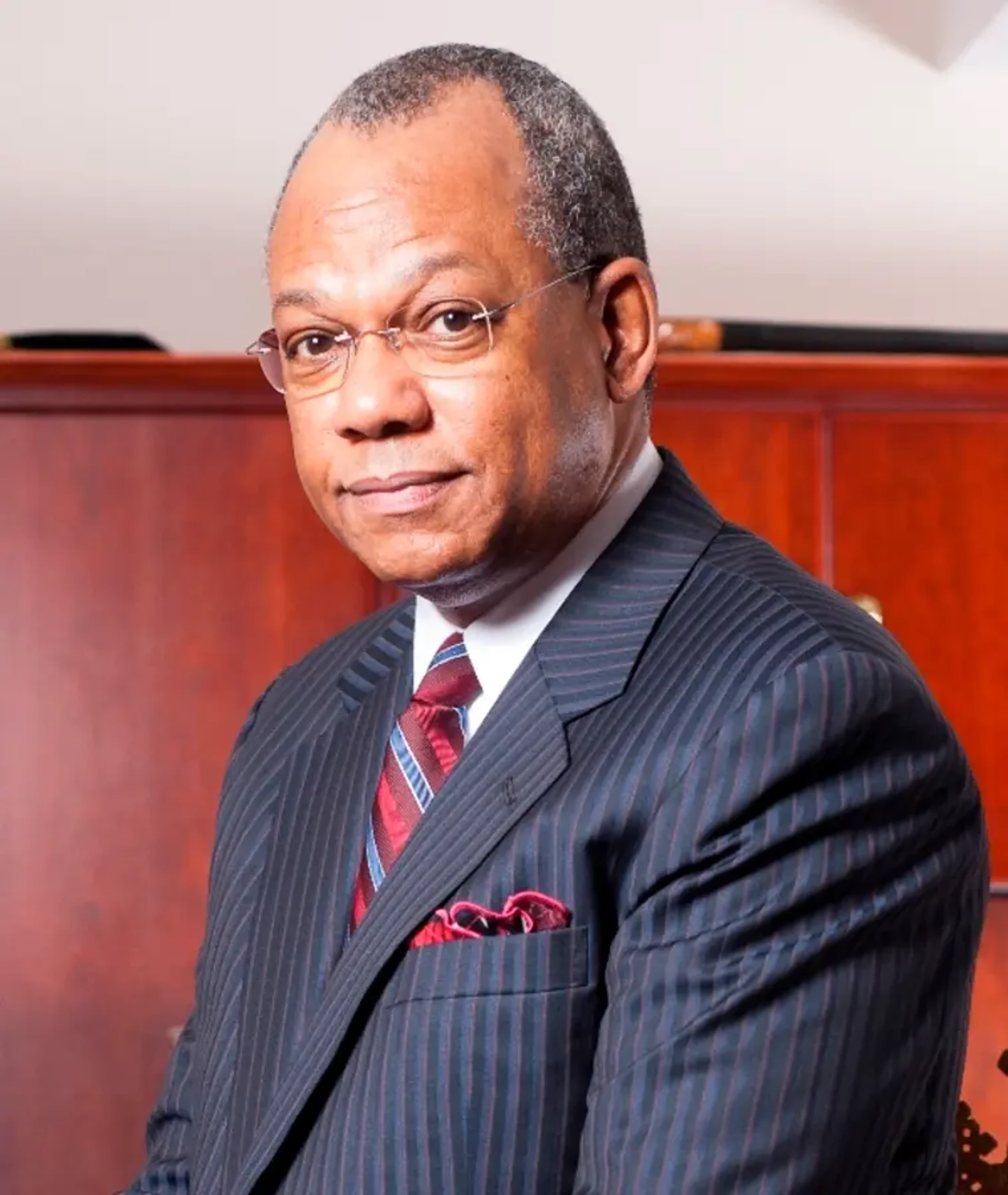 A professional headshot of a middle-aged Black man wearing glasses, a dark pinstripe suit, a red and blue tie, and a red pocket square.