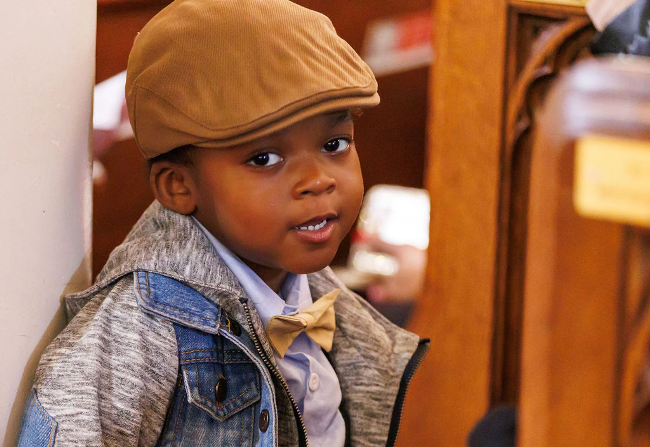 A young Black boy looks at the camera while wearing a tan flat cap, a matching bowtie, a light blue shirt, and a layered denim and grey jacket.