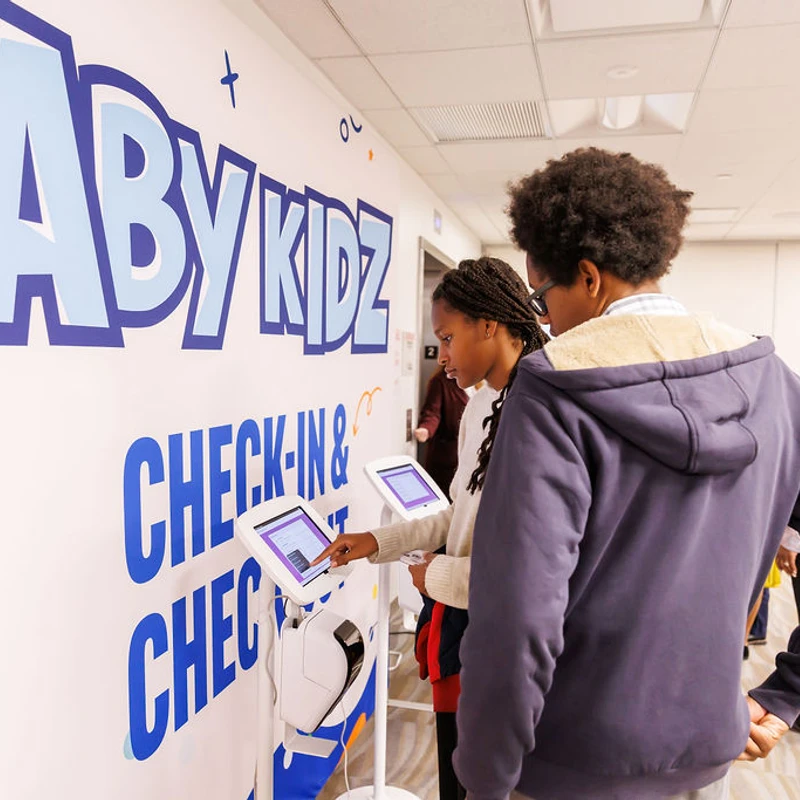 A young girl and boy stand in front of a wall with an 'ABY KIDZ CHECK-IN' sign, using a tablet on a stand to register for an event.