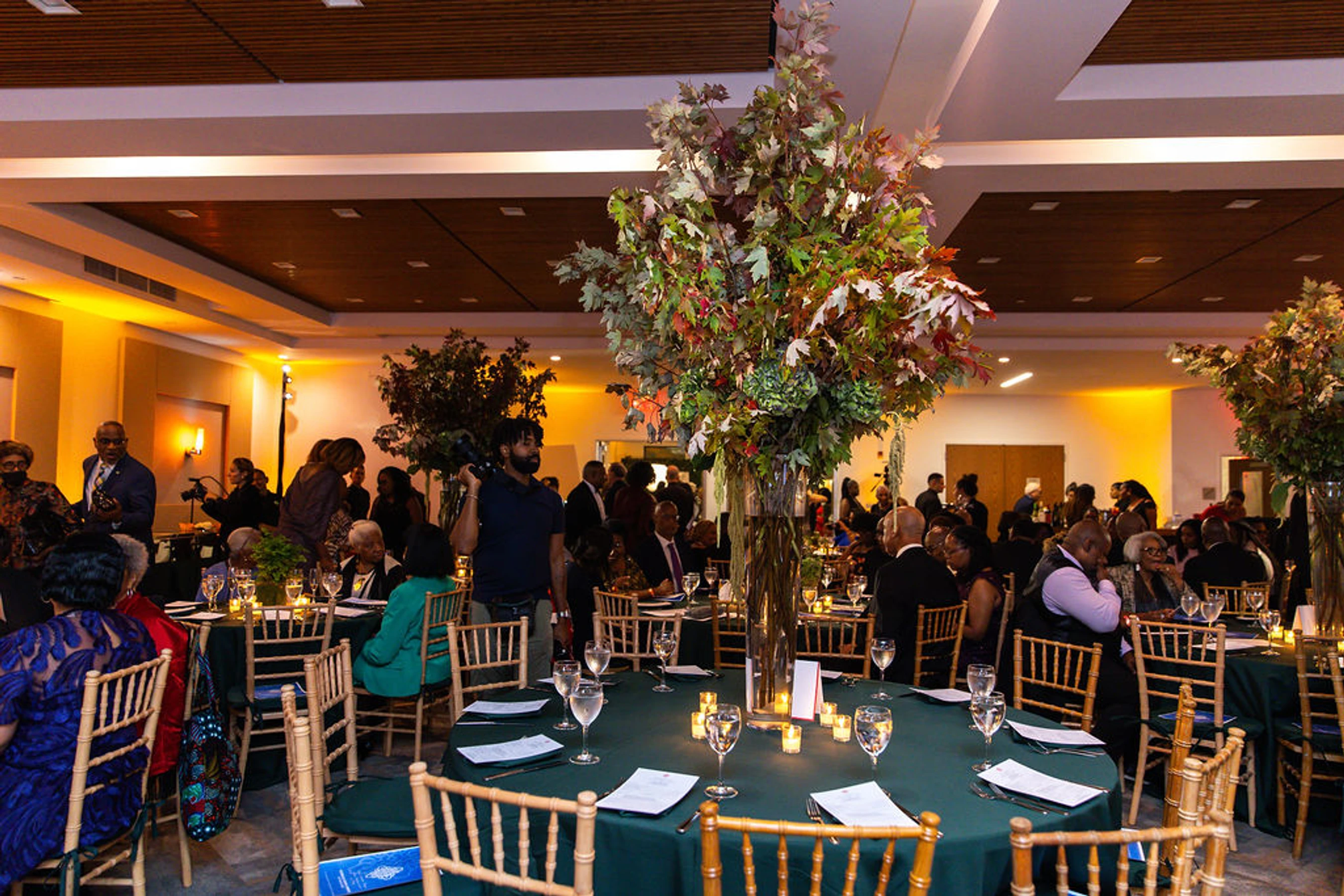 An event hall with guests seated and standing around tables with green cloths and large leafy centerpieces. A cameraman is filming the gathering.