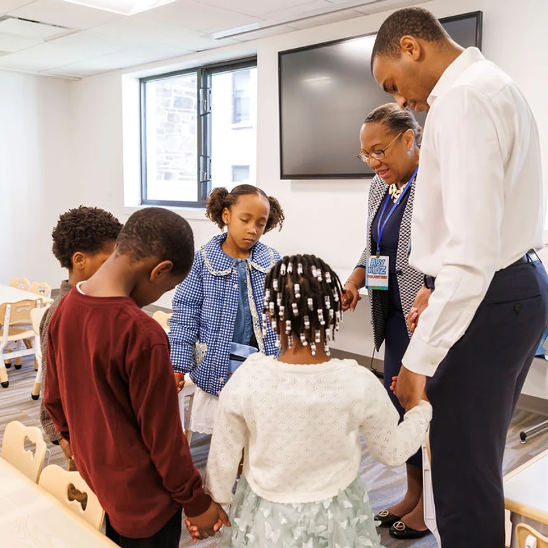 A group of young Black children and two Black adults hold hands in a circle, bowing their heads to pray in a brightly lit classroom.