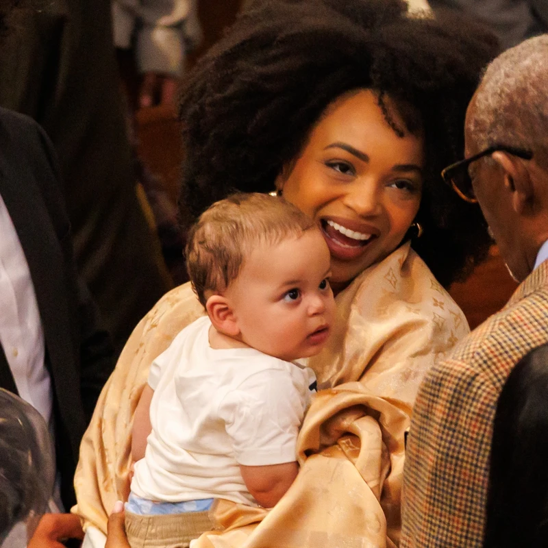 A smiling Black woman with voluminous curly hair holds a small baby boy while talking to an older man whose back is to the camera.