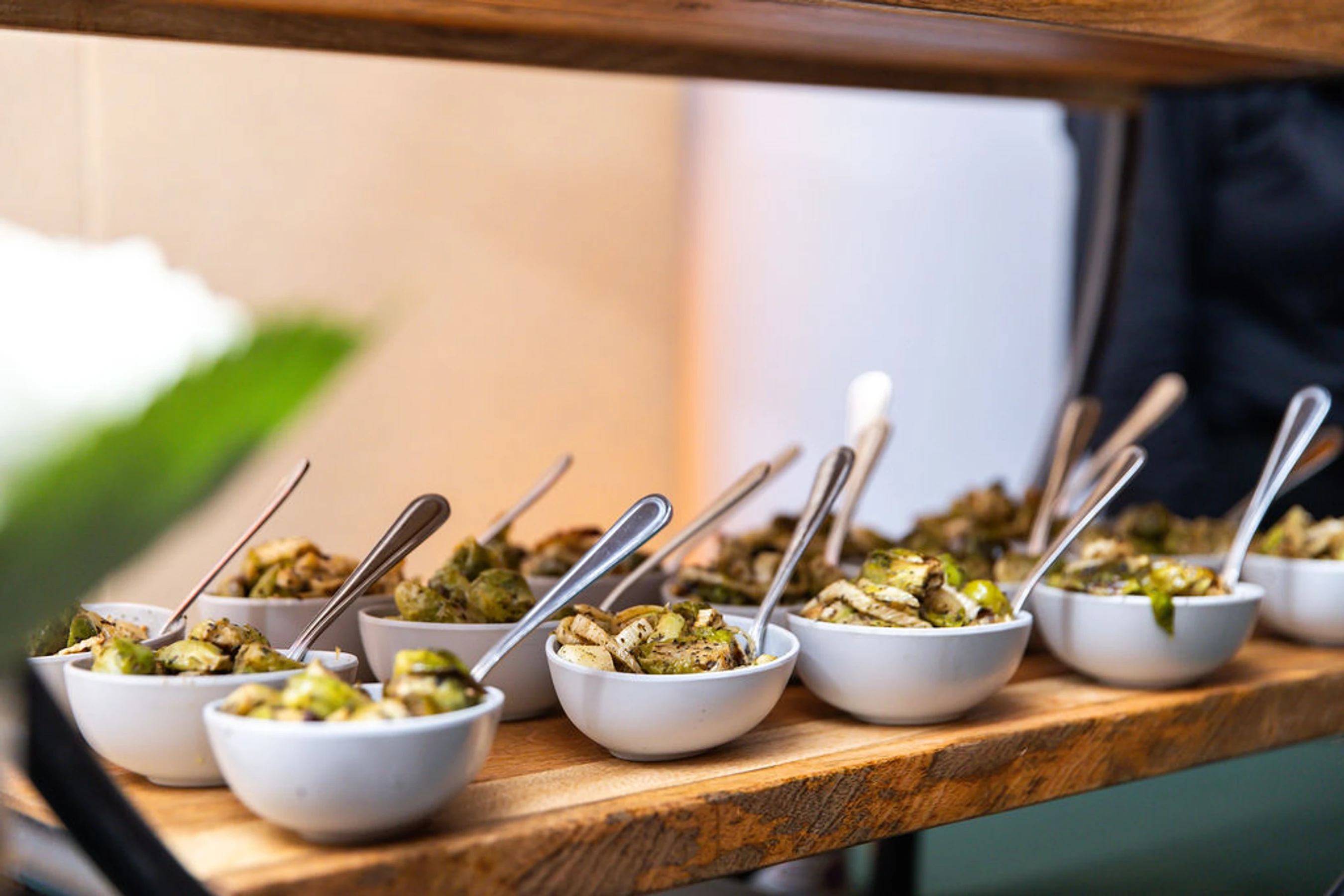 A close-up of small white bowls filled with roasted vegetables, likely brussels sprouts, each with a spoon, lined up on a rustic wooden serving tray.