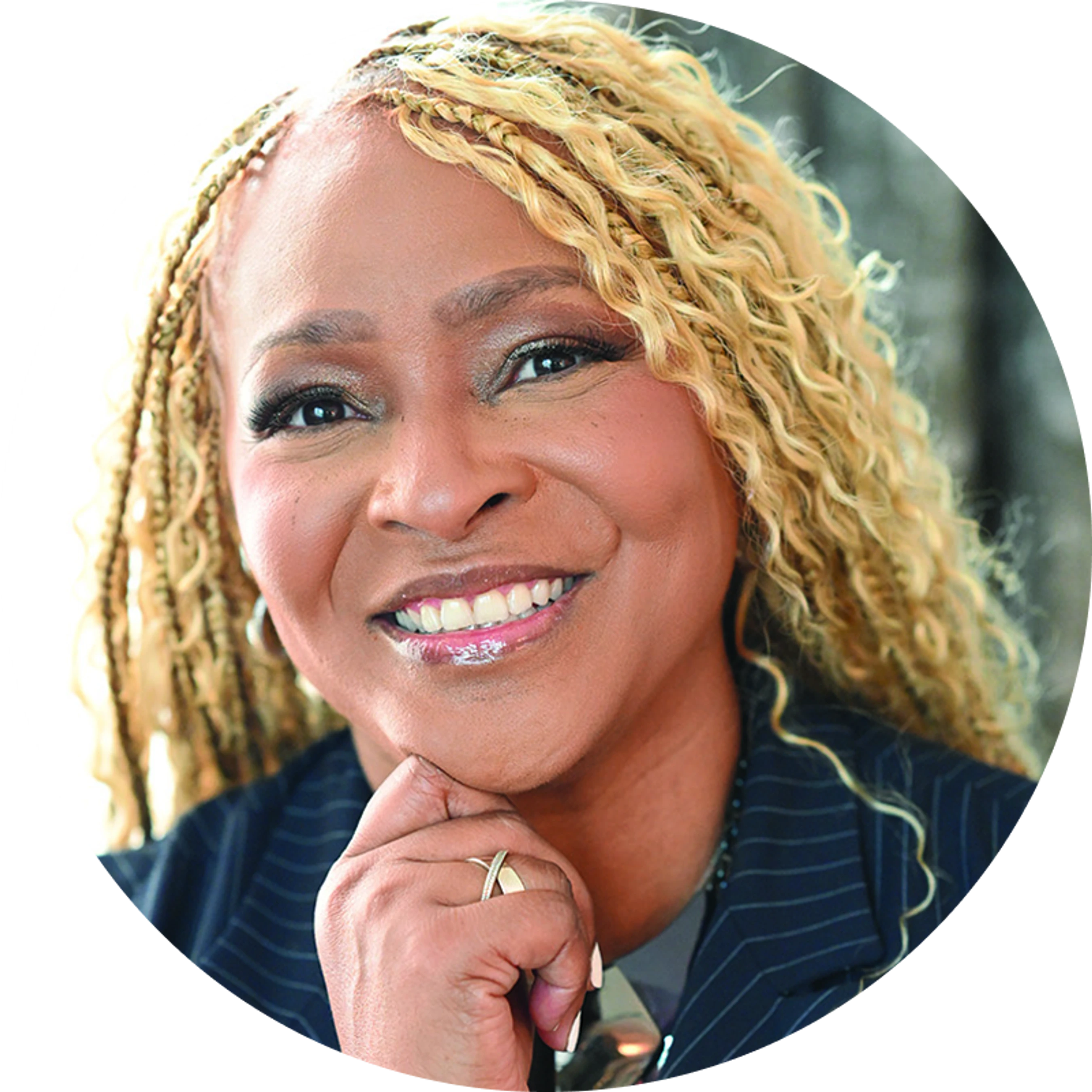 A close-up, circular headshot of a smiling Black woman with long, blonde braids and curls. She is wearing a pinstripe blazer and resting her chin on her hand.