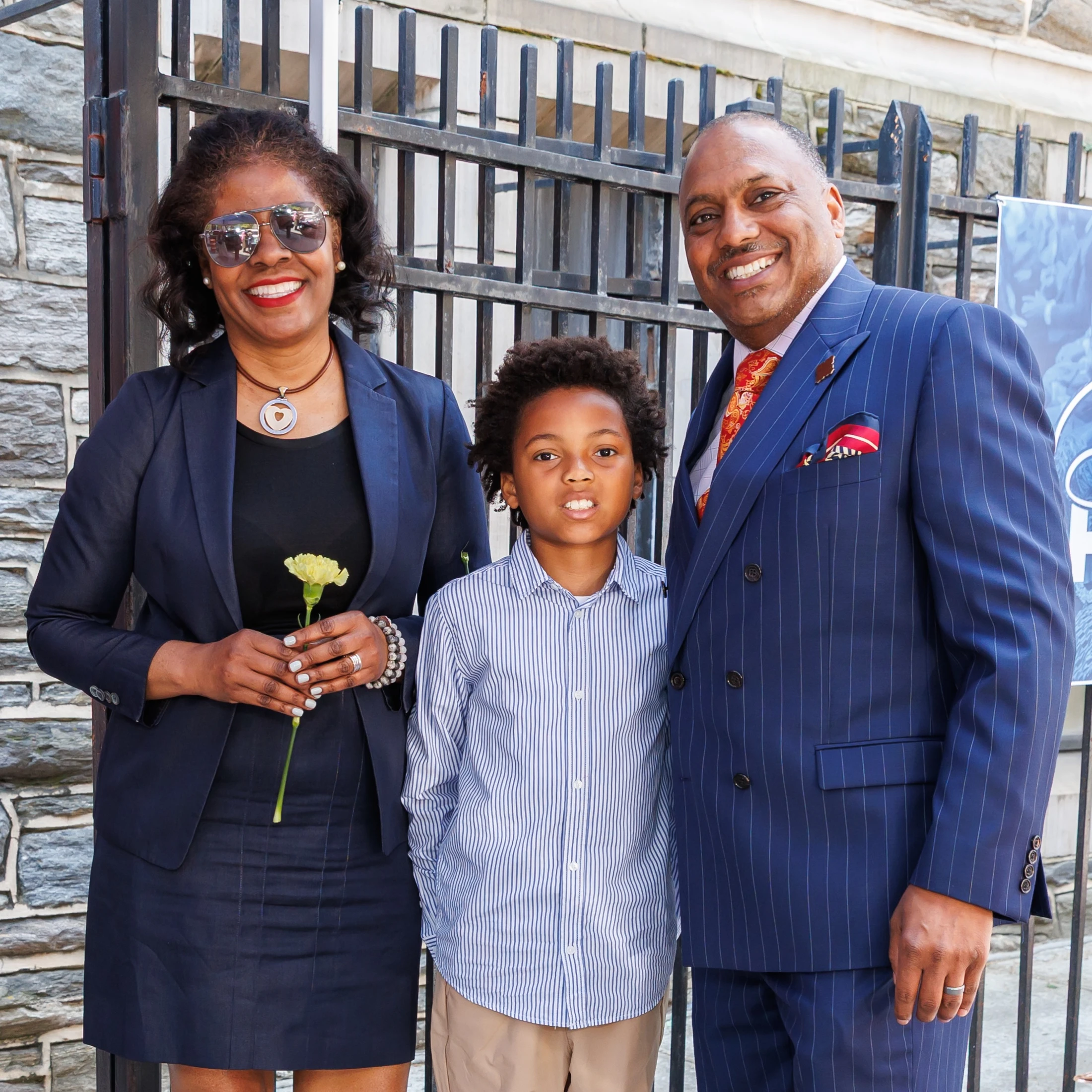 A Black family of three—a woman in a suit, a man in a pinstripe suit, and a young boy—smile for a photo while standing in front of a metal gate.