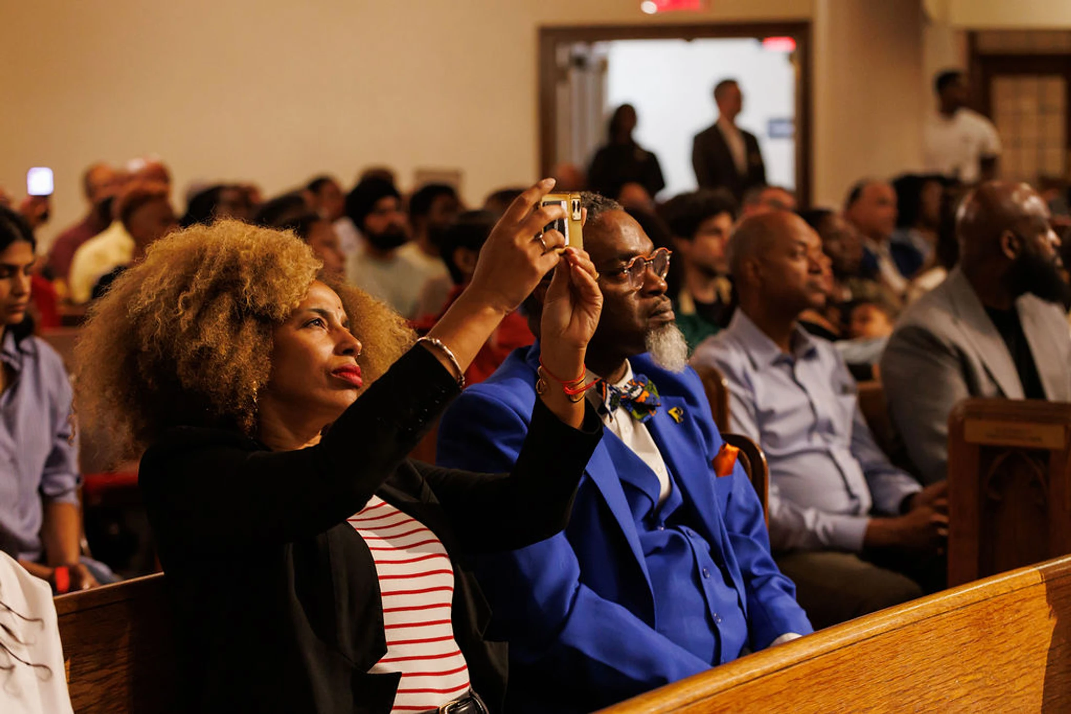 A woman with blonde curly hair holds a smartphone to take a photo while sitting in a pew. Next to her, a man in a blue suit watches the event.