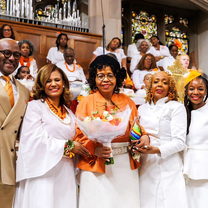 A group of five Black people smiling for a photo in a church. The woman in the center holds a bouquet, surrounded by a congregation dressed in white and orange.