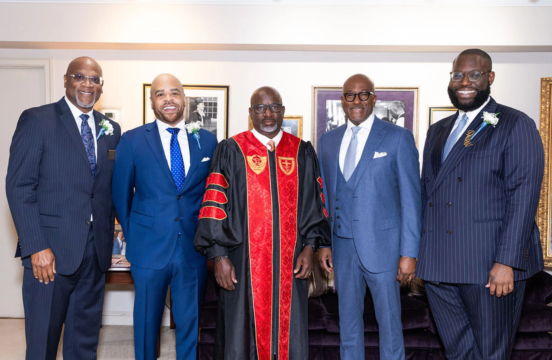 Five smiling Black men pose together indoors. Four wear suits and one in the center wears a black and red academic or clerical robe. They all wear boutonnieres.