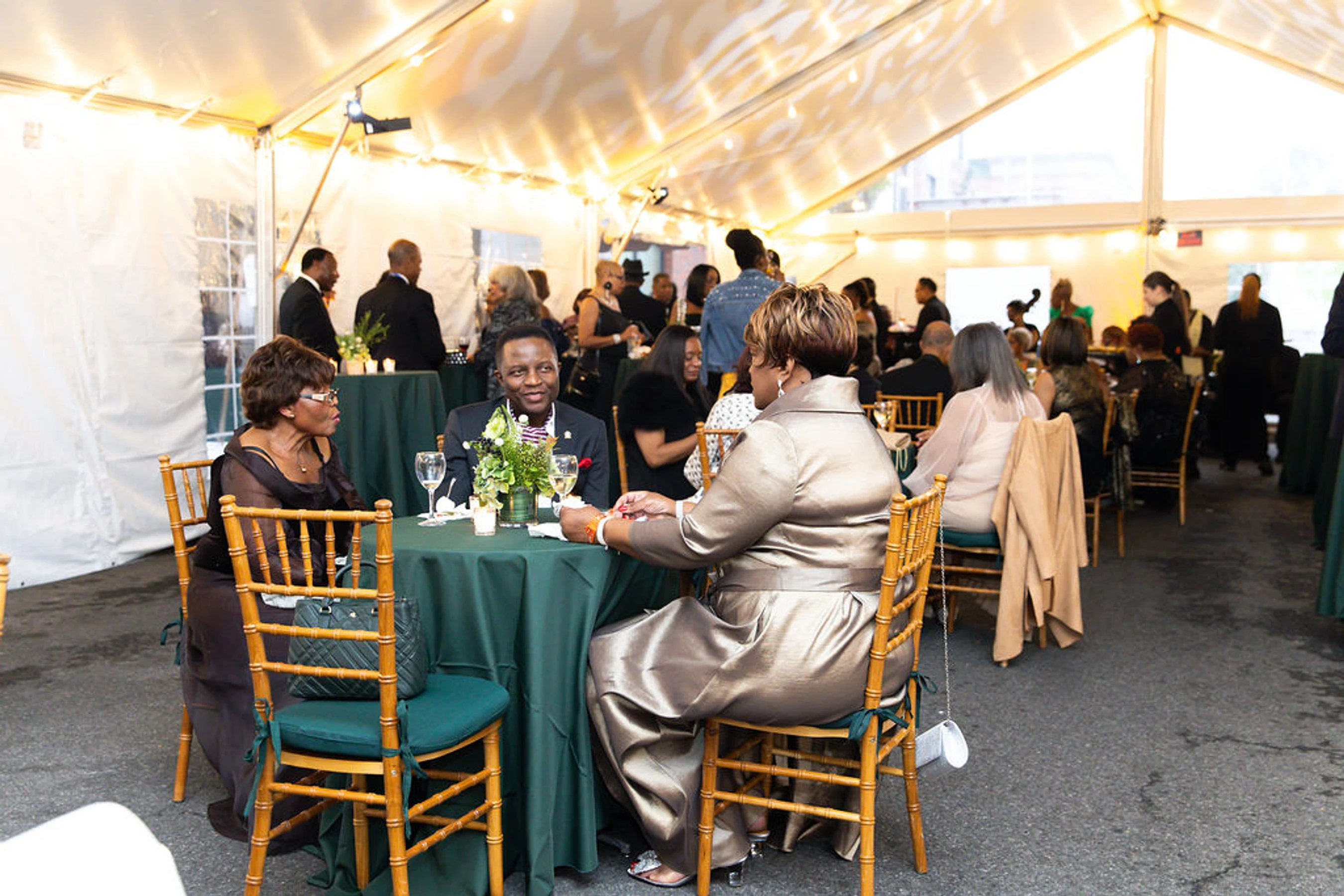 Three formally dressed guests chat at a green-covered table inside a large, lit tent during a gala, with other attendees mingling in the background.