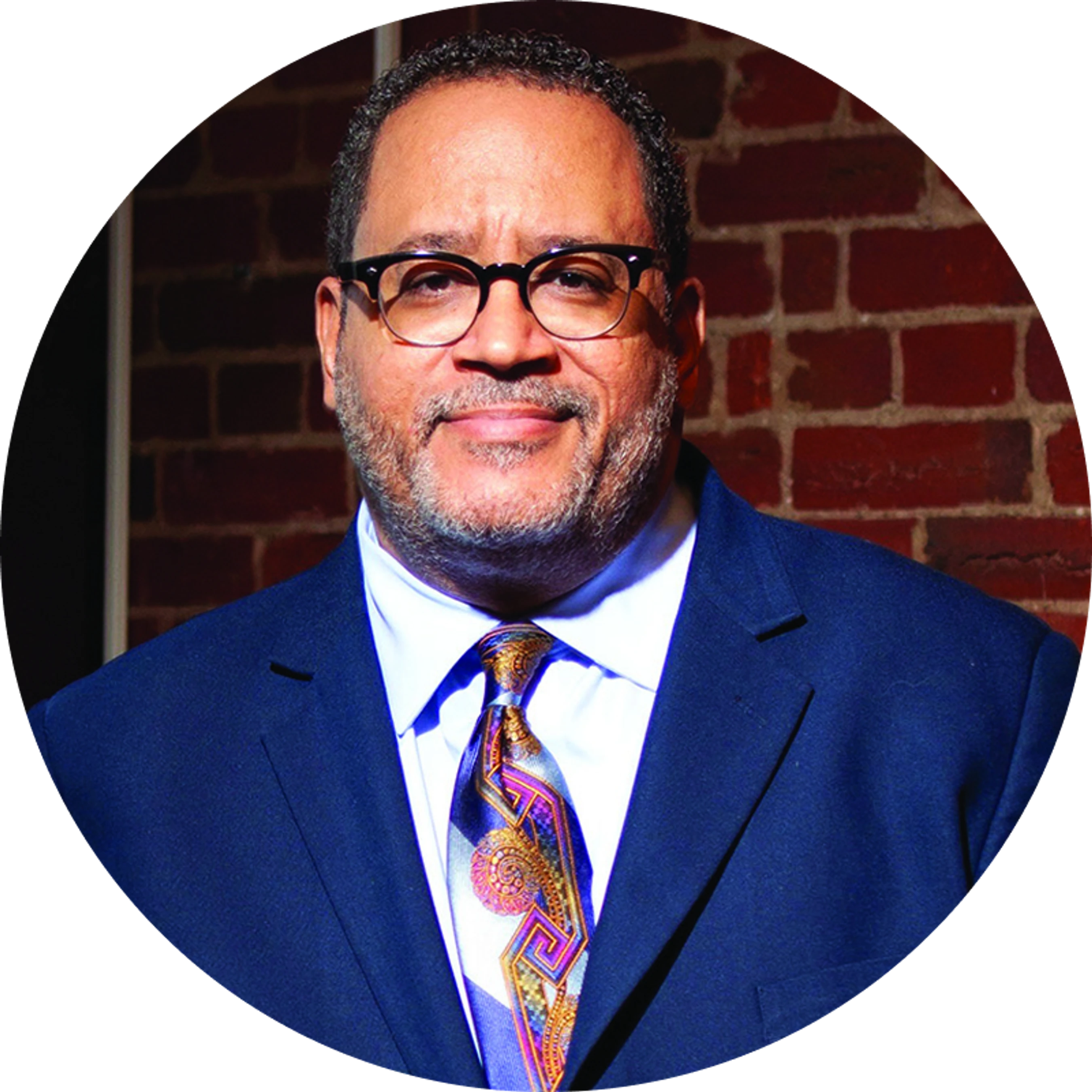 A circular headshot of a smiling man with glasses, wearing a blue suit and a colorful patterned tie, in front of a brick wall.