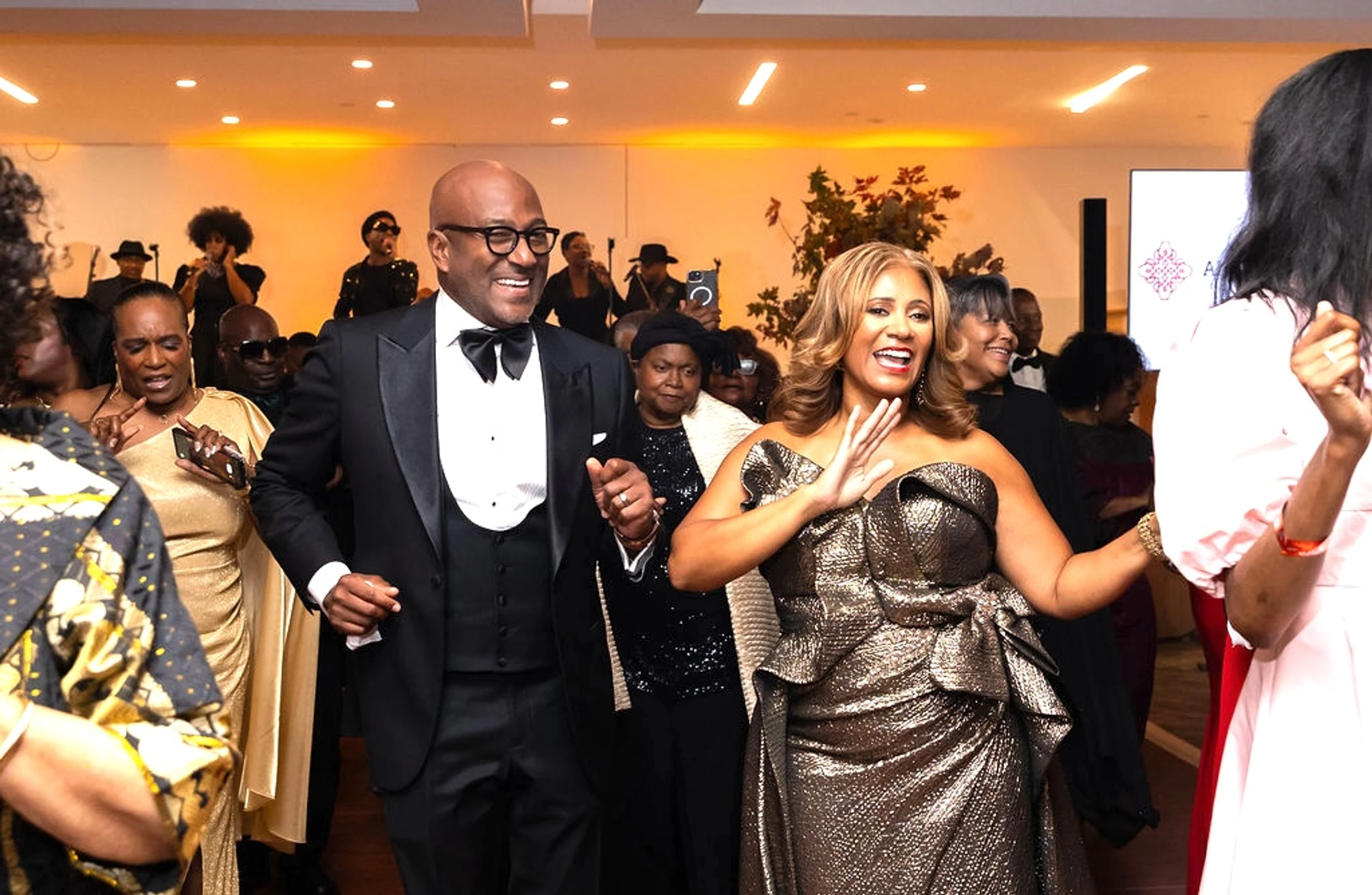 A man in a tuxedo and a woman in a metallic gown smile and dance together in a crowd at an indoor event, with other formally dressed guests around them.