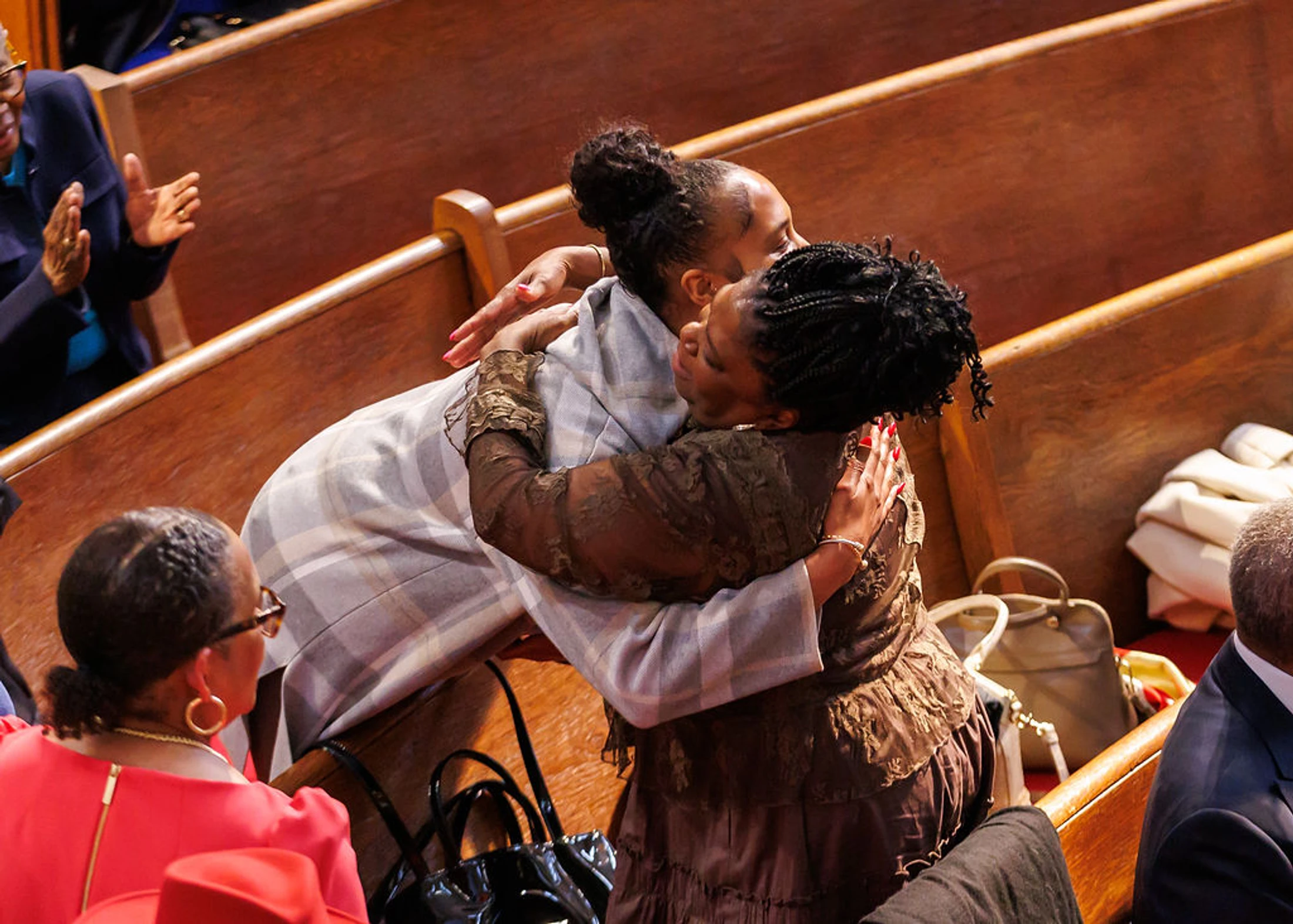 Two Black women share an emotional hug in a wooden church pew. Other members of the congregation are seated around them, some clapping in the background.