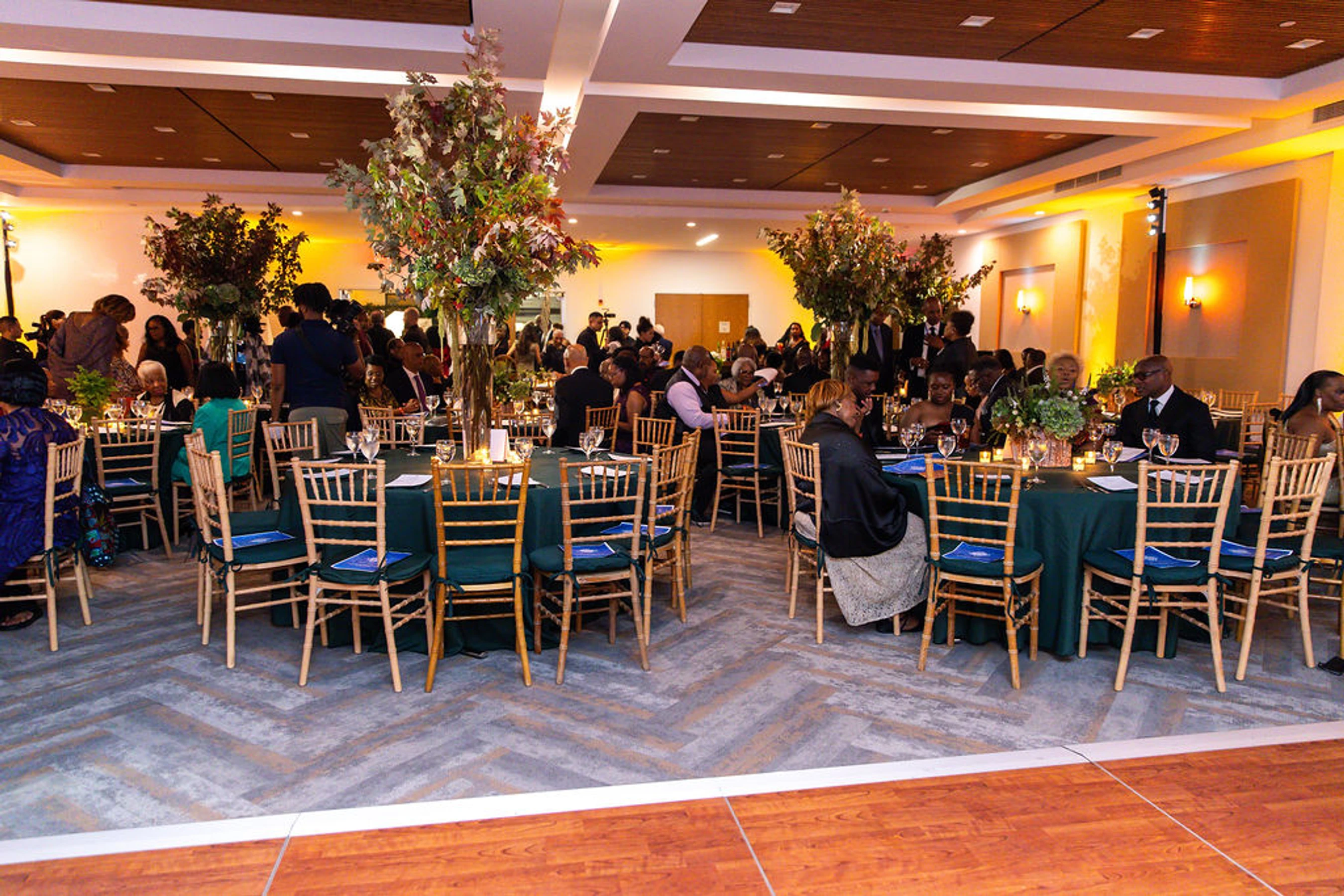 A banquet hall filled with guests at round tables with green tablecloths and tall floral centerpieces. The room has warm lighting and elegant decor.