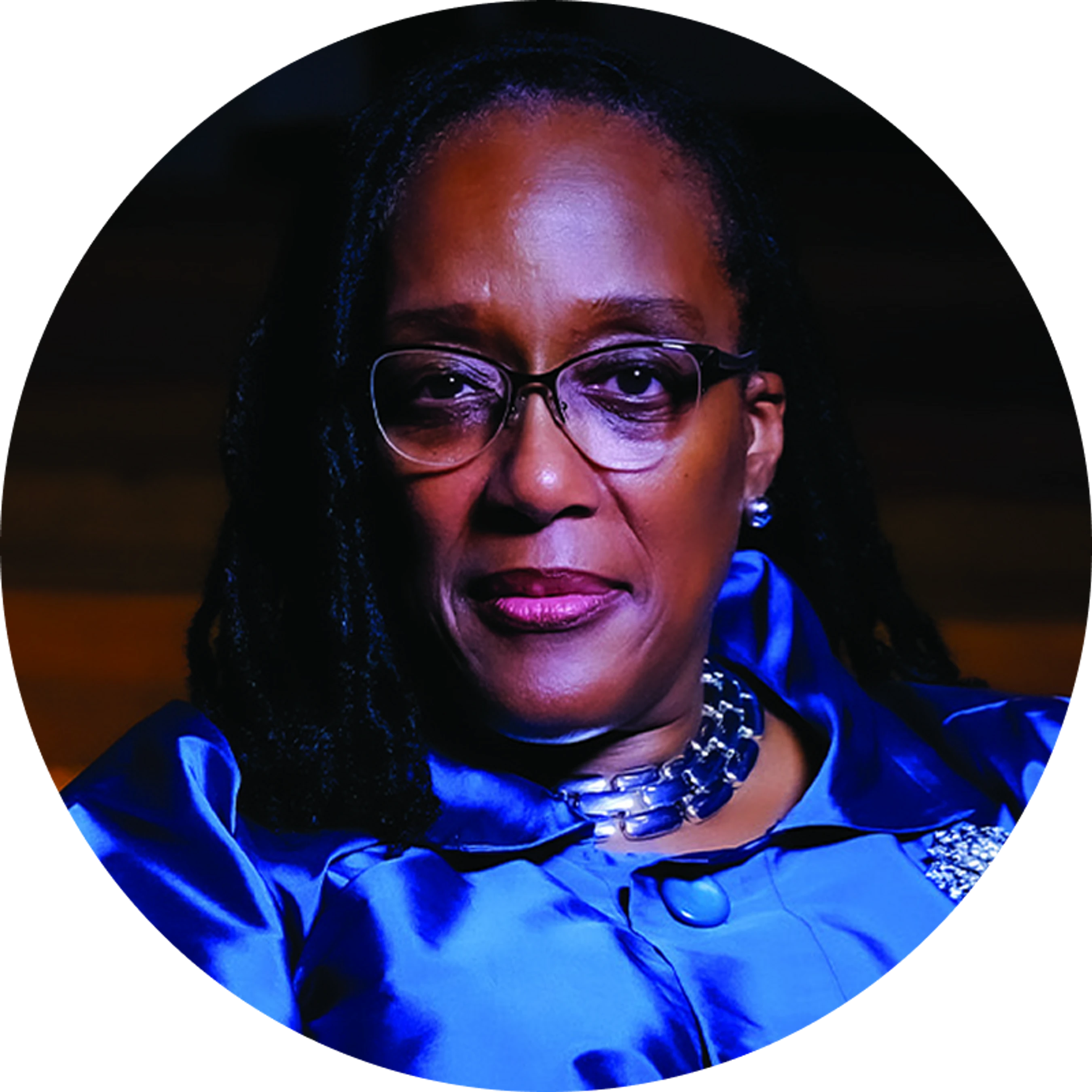 A circular headshot of a Black woman with dreadlocks and glasses, wearing a vibrant blue satin jacket and a chunky silver necklace, looking at the camera.