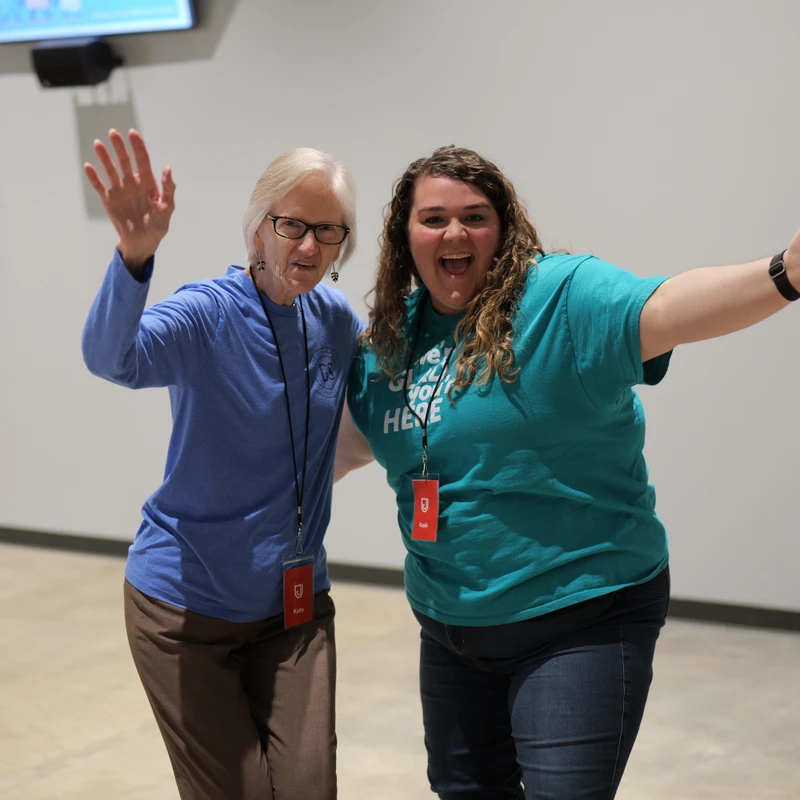 An older woman in a blue shirt waves and a younger woman in a teal shirt holds her arms out, both smiling happily for the camera in a large room.